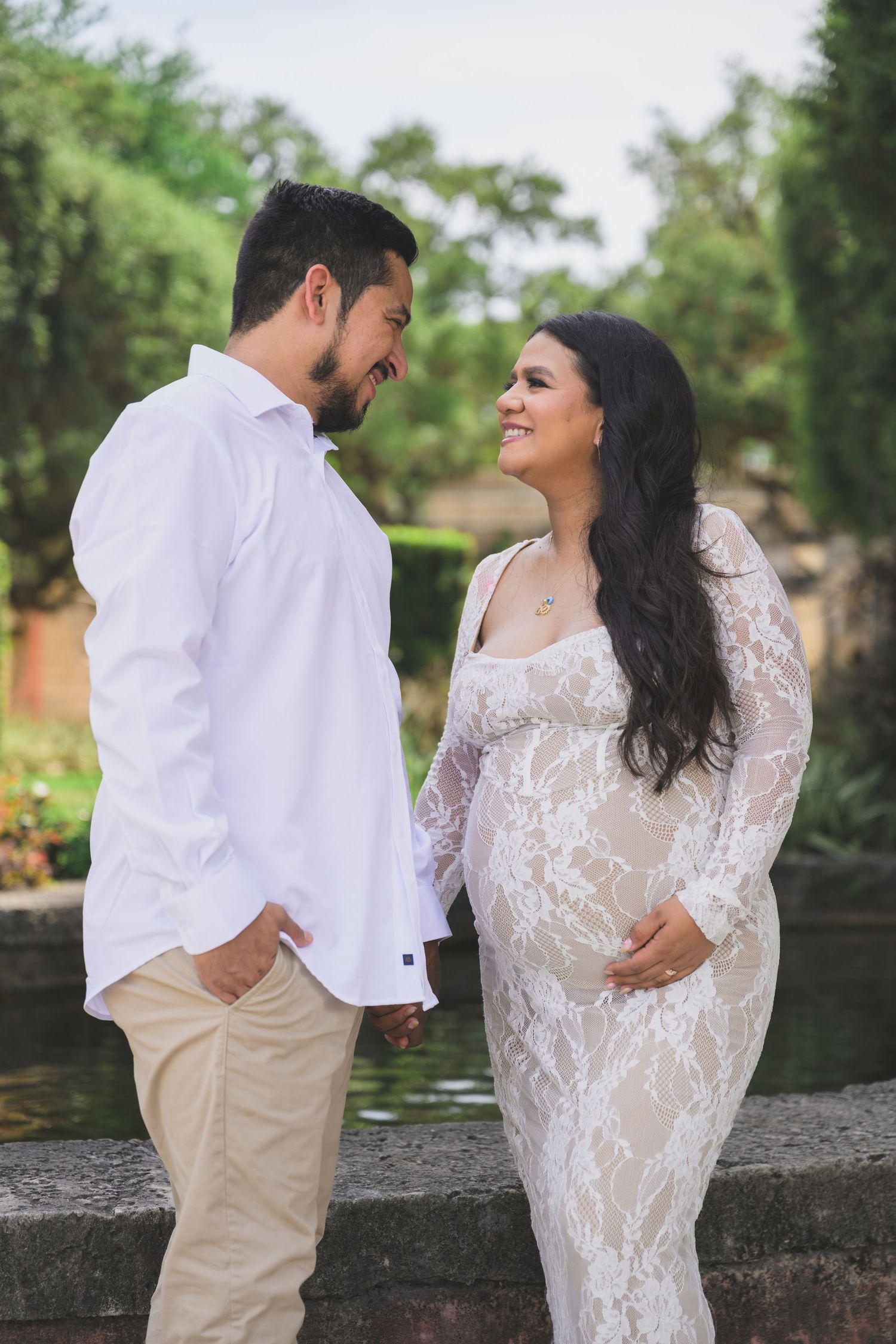 Couple sharing a tender moment outdoors, pregnant woman in white lace gown holding baby bump, man in white shirt and khakis.