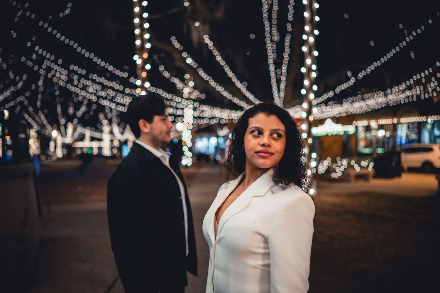 Couple posing at night under sparkling string lights, woman in white blazer looking back at camera.