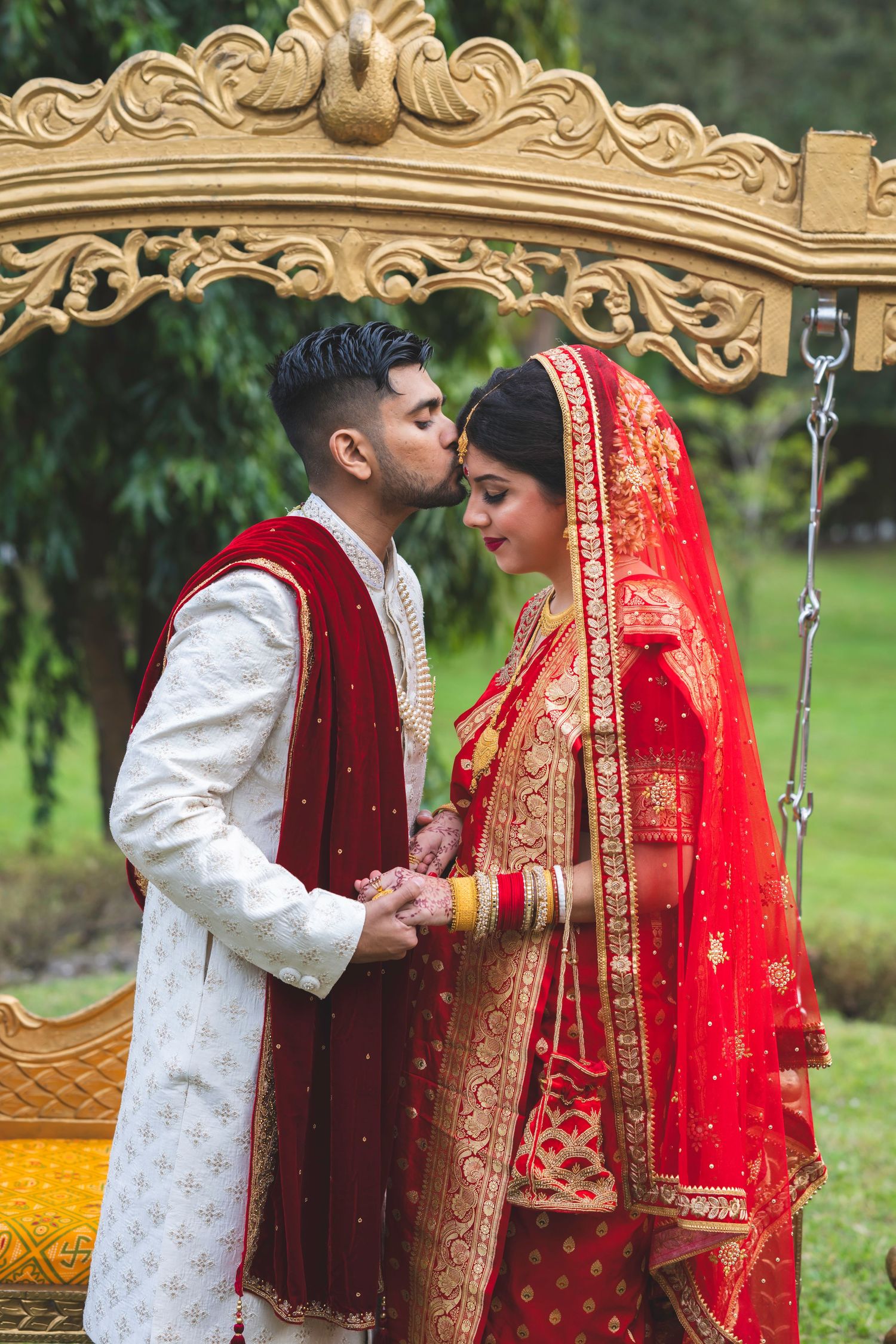 Indian groom in white sherwani kisses bride in red and gold lehenga under ornate golden mandap outdoors.