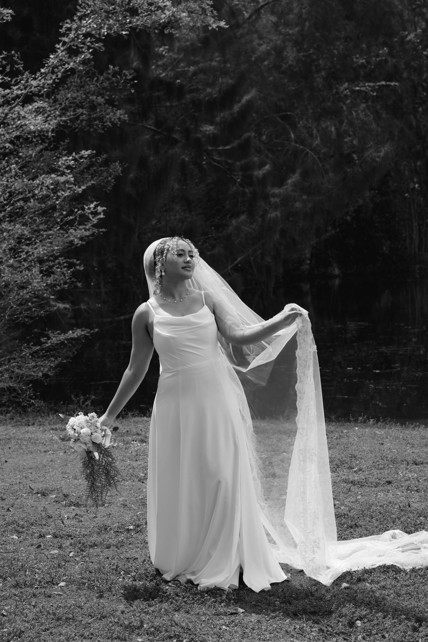 Black and white portrait of a bride in a flowing gown holding bouquet while her veil blows in wind.