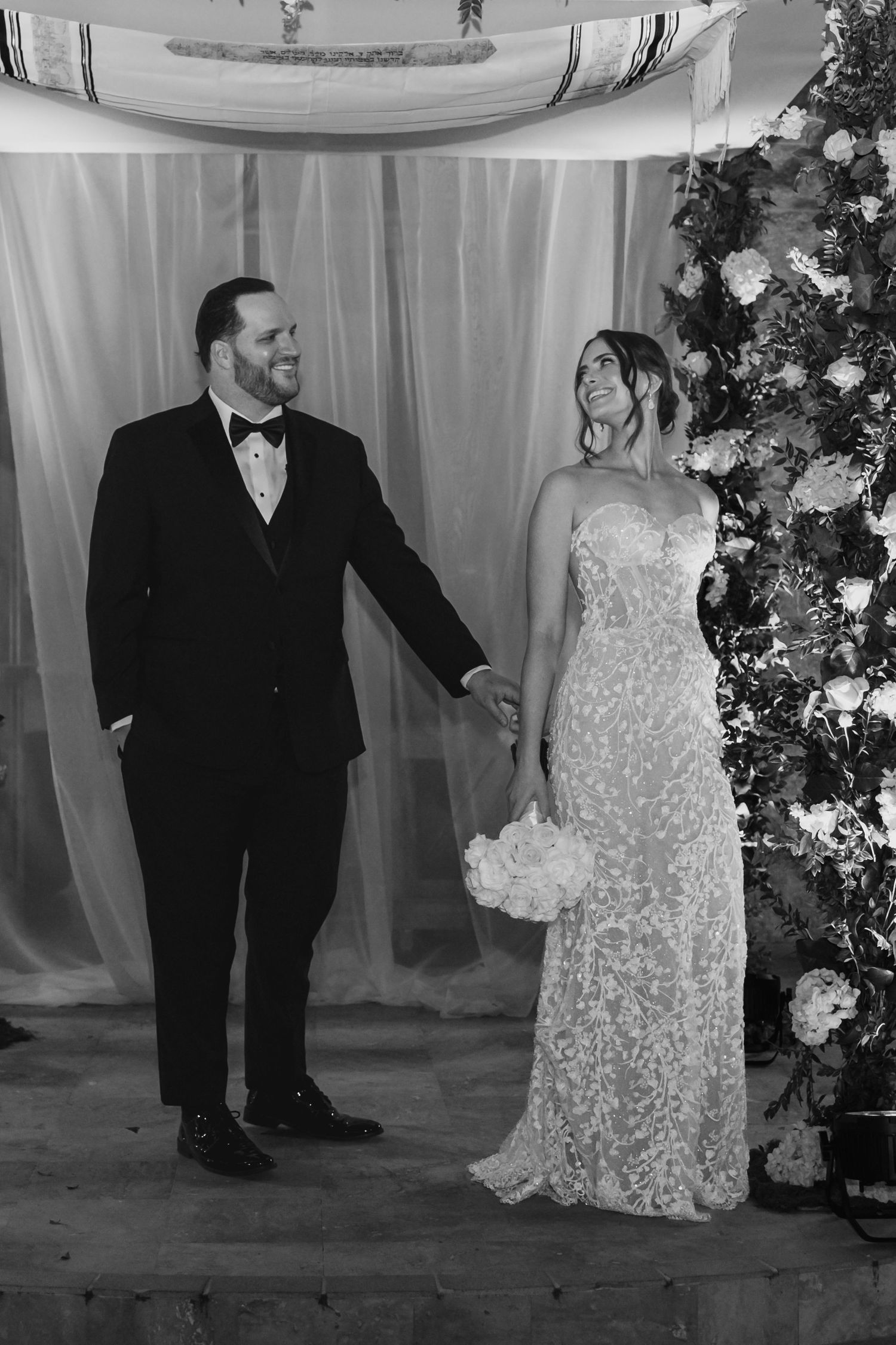 Black and white photo of bride and groom holding hands at floral wedding altar.