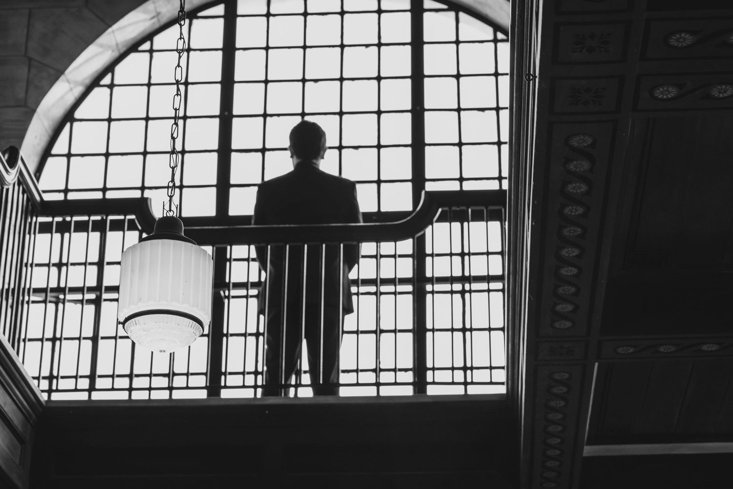 Silhouetted man standing at a balcony railing before a large arched window in a grand building, black and white photo.
