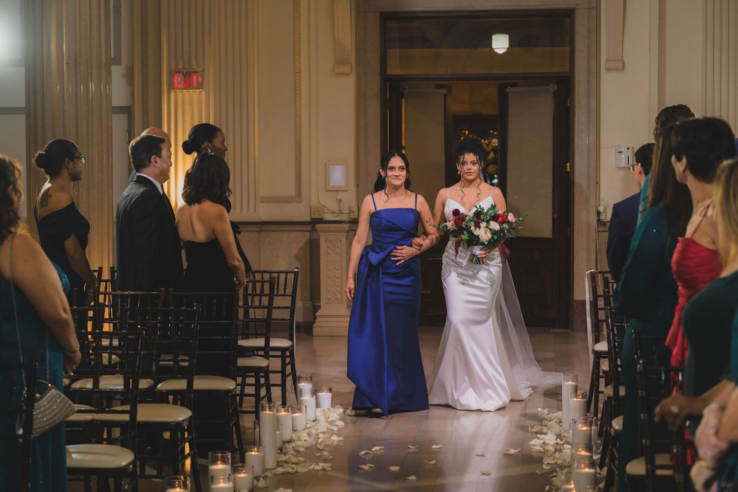Bride in white gown walks down aisle with bridesmaid in royal blue dress at elegant indoor wedding ceremony.