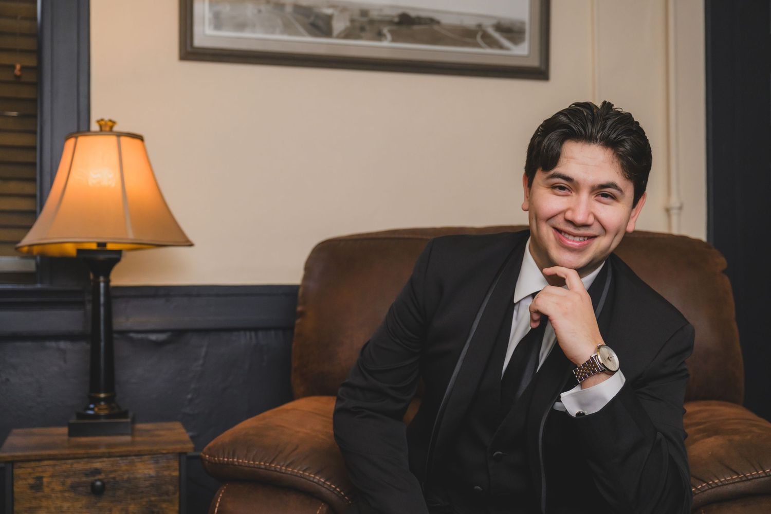 Smiling young man in black suit sitting relaxed on leather chair beside a lit table lamp in elegant room.