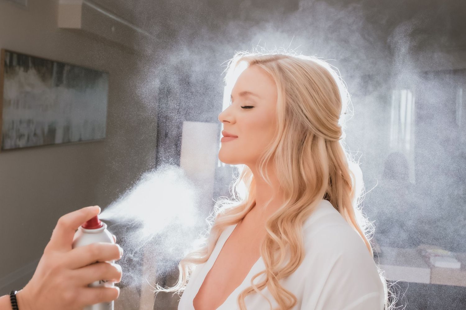 Blonde bride smiling with eyes closed while getting hairspray applied during wedding preparation.