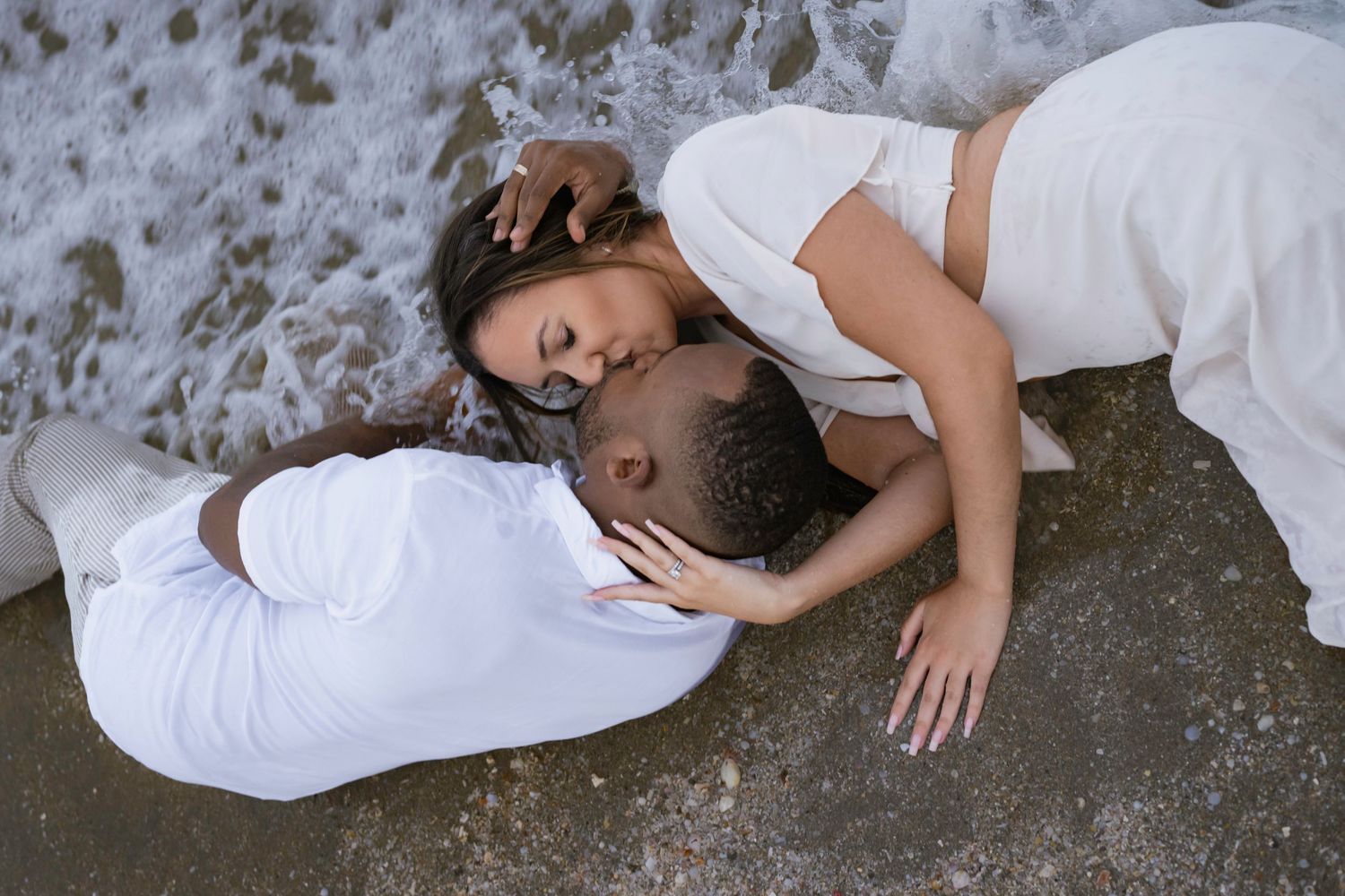 Romantic couple in white outfits lying on wet rocks as ocean waves crash around them.