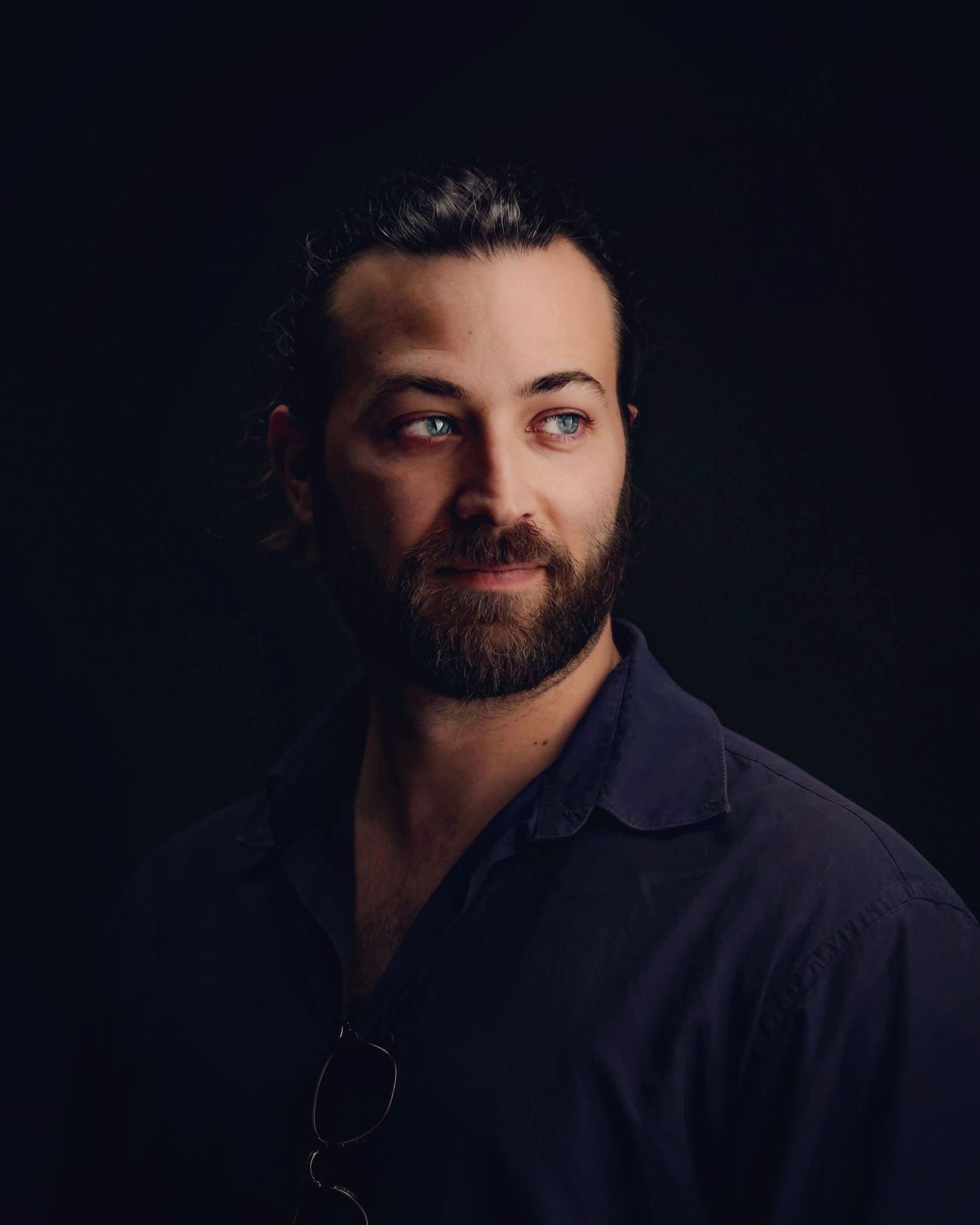 Professional portrait of a bearded man with blue eyes in a dark shirt against a black background.