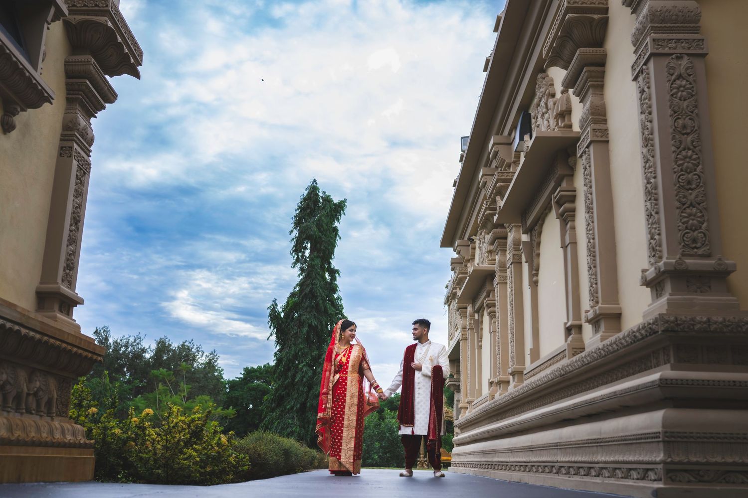 Indian bride and groom in traditional red and white attire walk between ornate temple columns under blue sky.