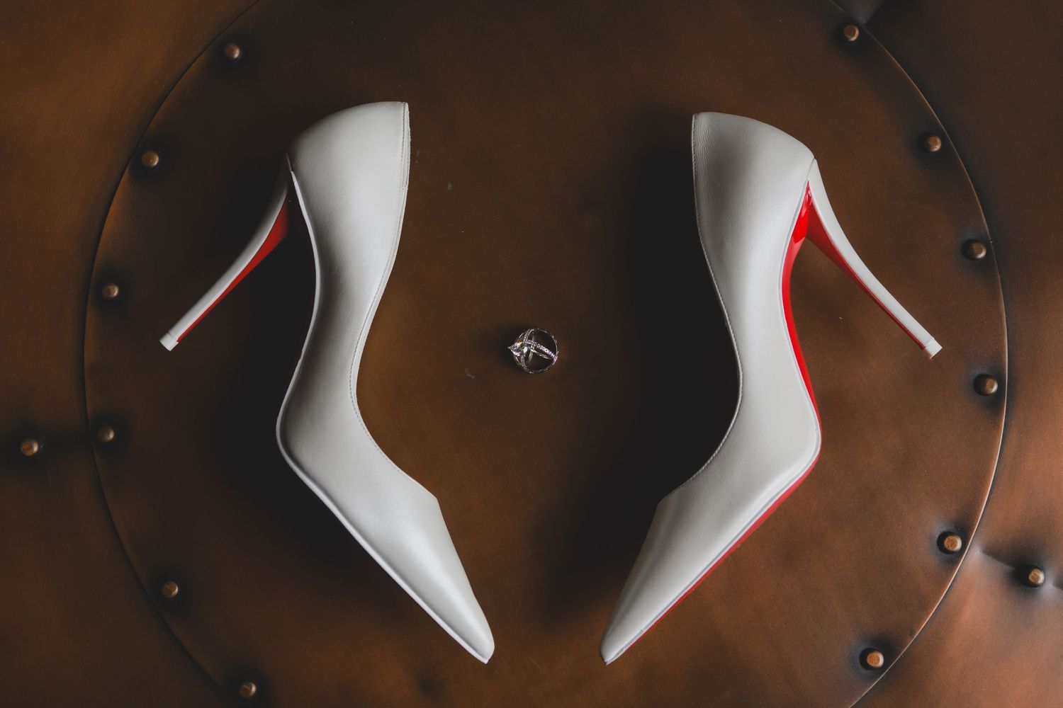 White Christian Louboutin wedding heels with red soles displayed on leather surface with diamond ring.