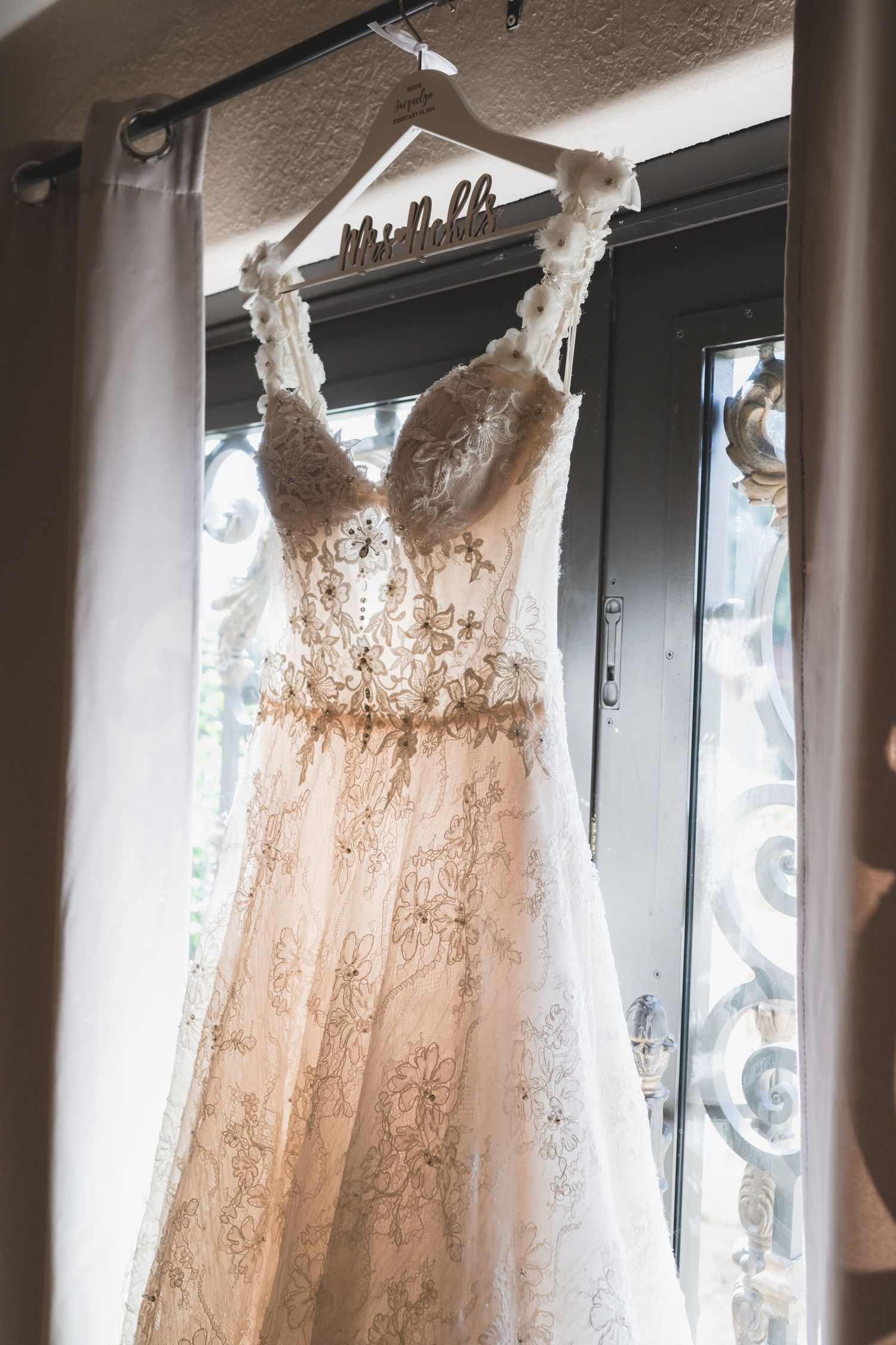 Intricate lace and beaded A-line wedding dress hanging on personalized hanger in window light.