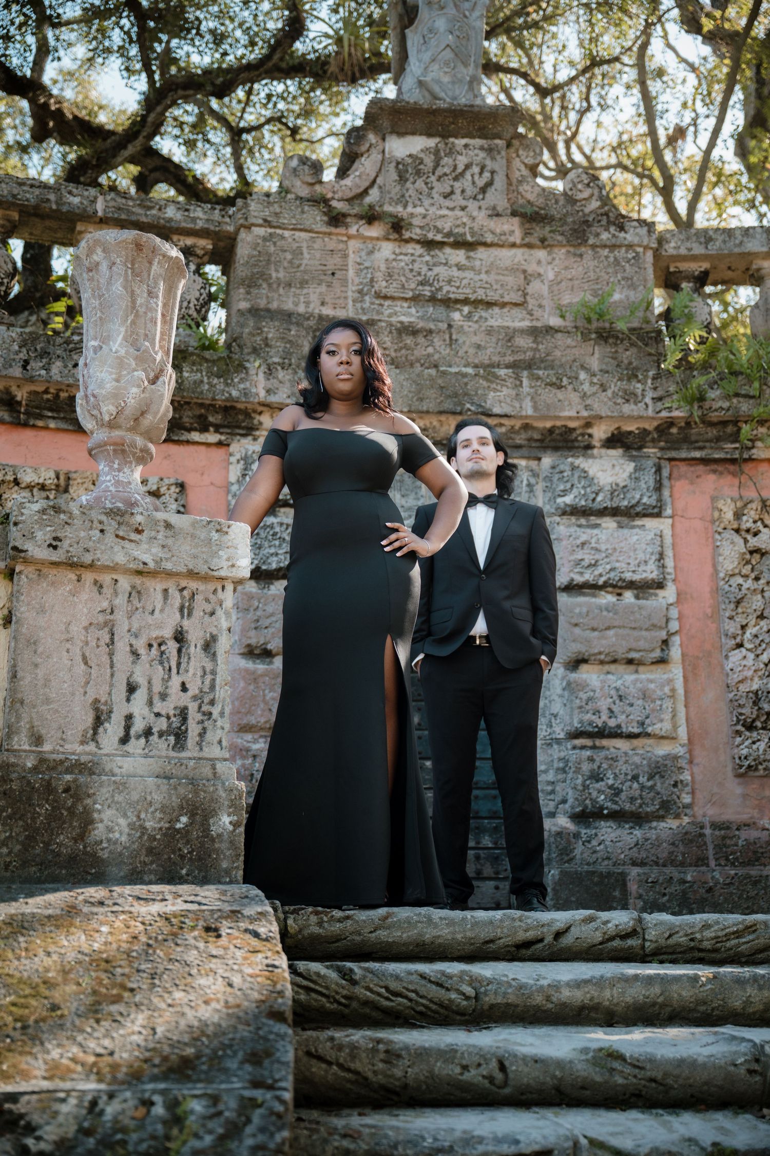 Elegant couple in matching black formal attire posing on stone steps at Vizcaya Museum.