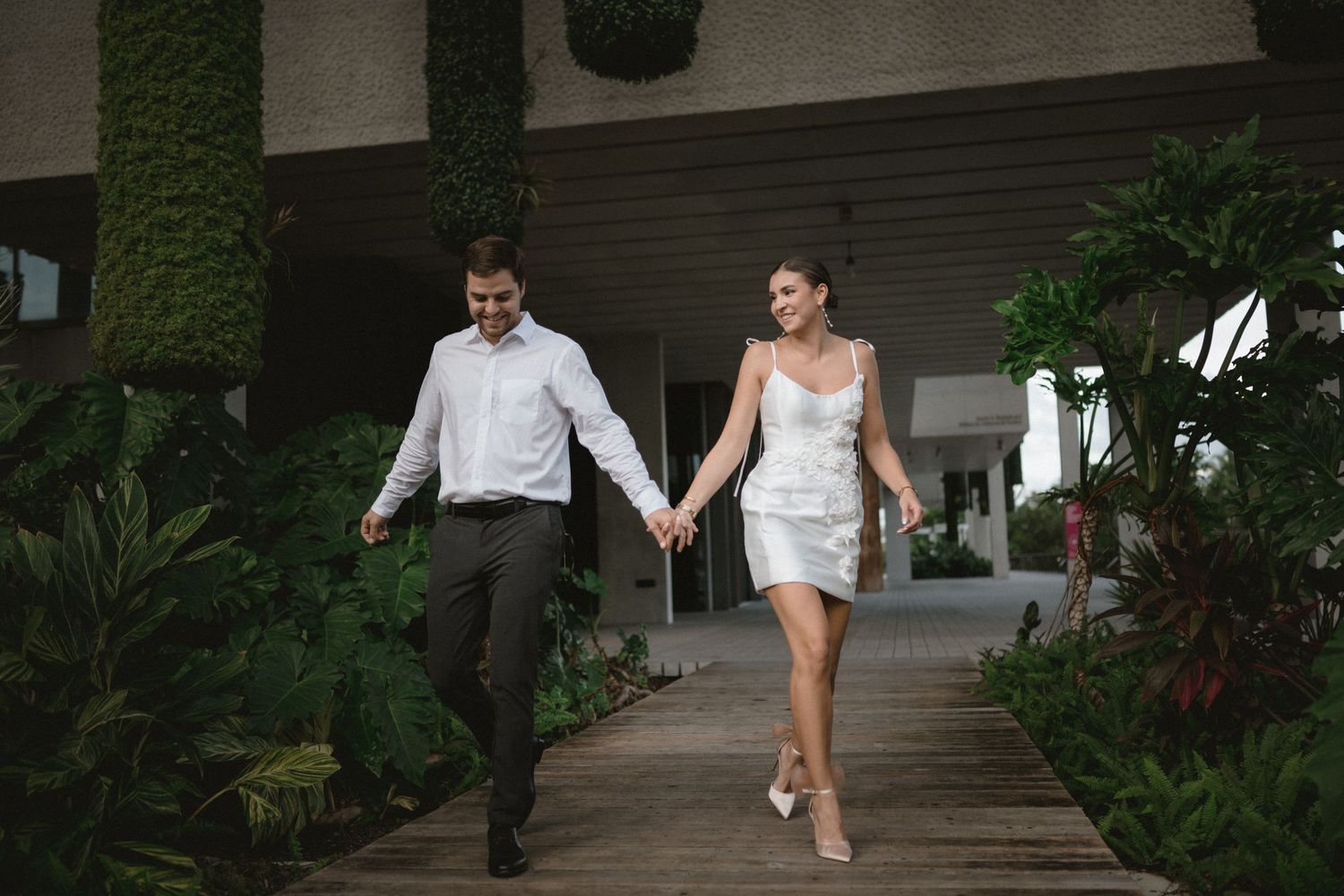 Elegant couple holding hands walking along a wooden path surrounded by lush tropical greenery.