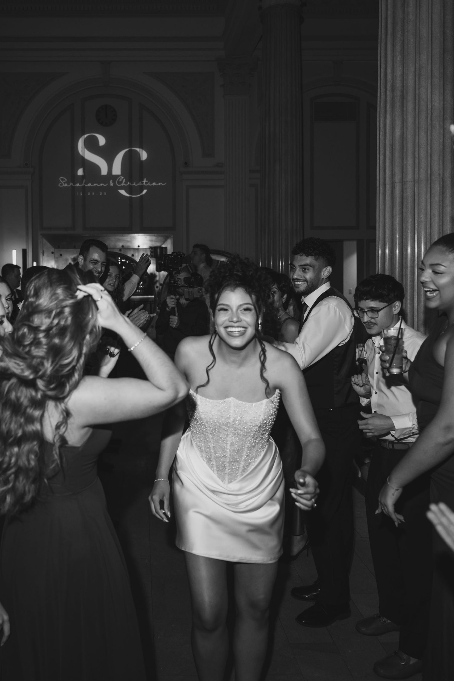 Bride in short white dress dancing joyfully with guests at wedding reception in black and white photo.