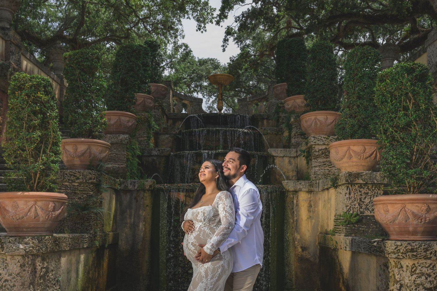Pregnant couple poses romantically before a lush garden waterfall surrounded by terracotta urns and greenery.