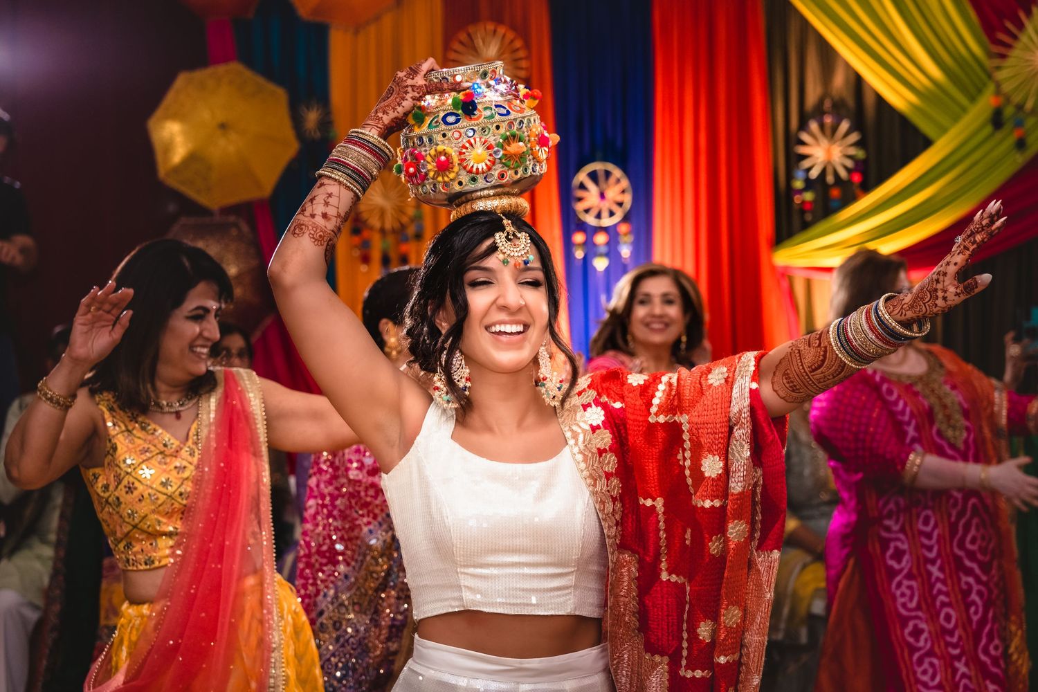 Joyful Indian bride dancing and holding a decorated pot above her head at a vibrant Mehndi celebration.
