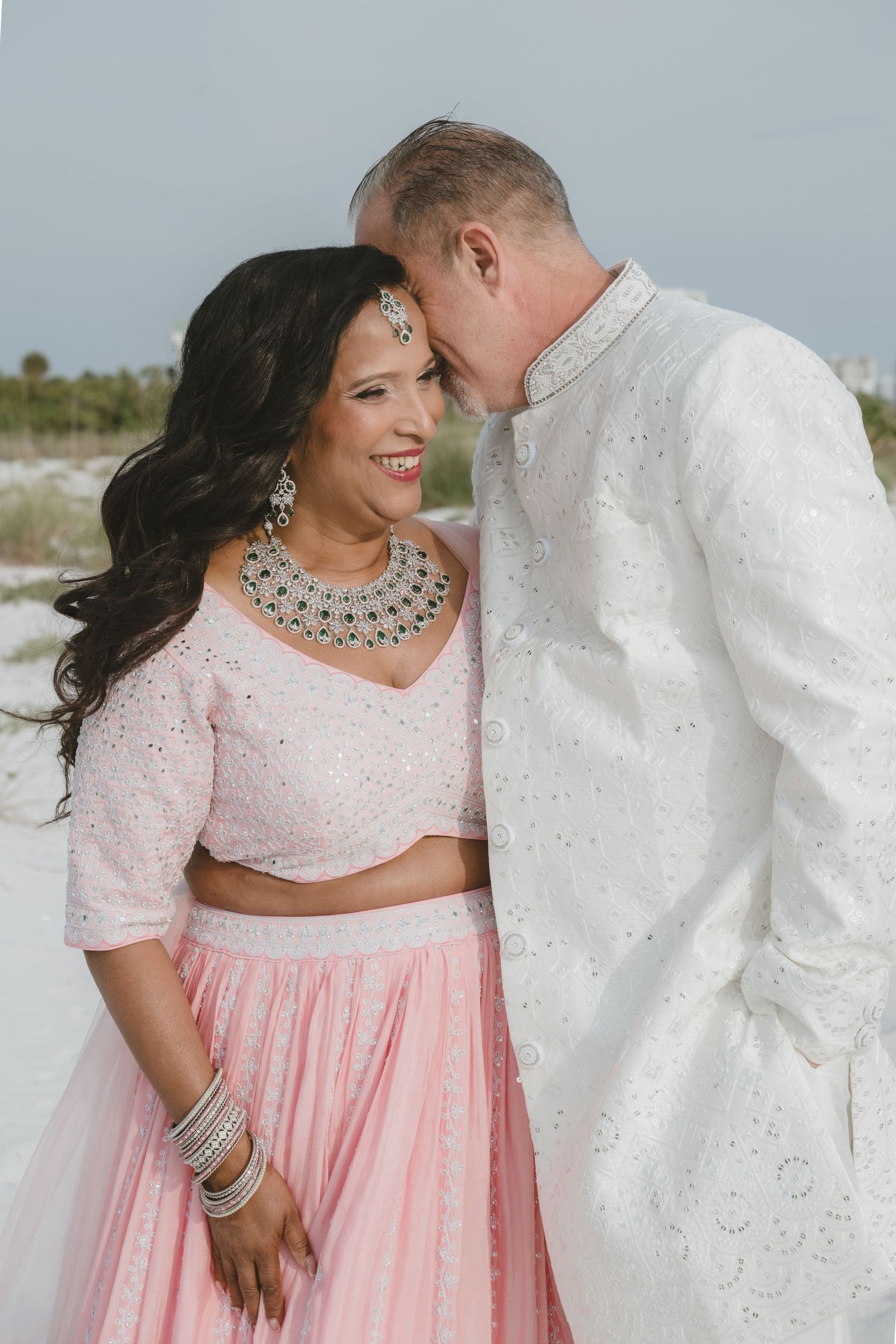 Bride in pink lehenga with silver jewelry smiles as groom in white sherwani leans close on a beach.