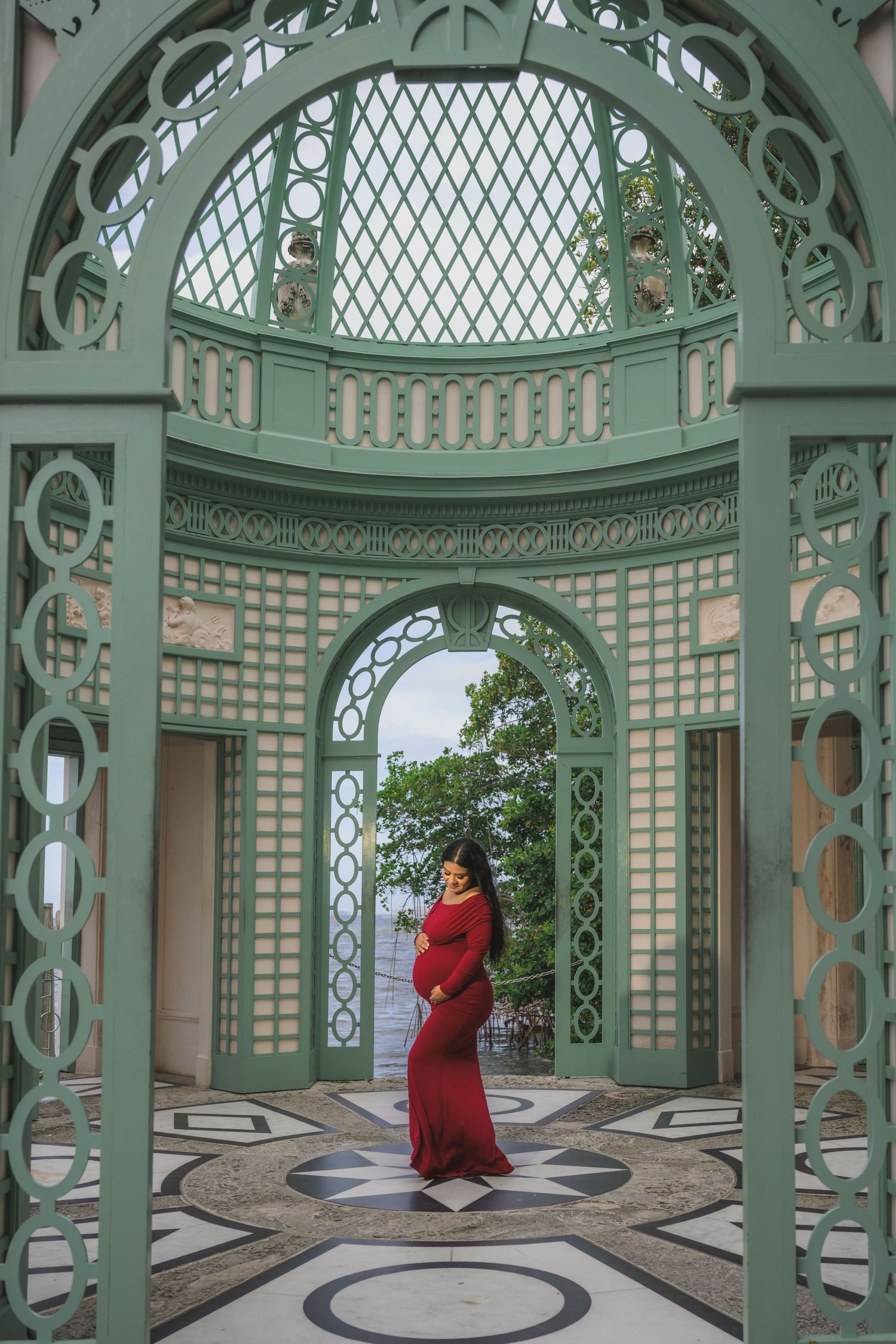 Pregnant woman in red gown poses inside ornate green lattice pavilion with ocean view.