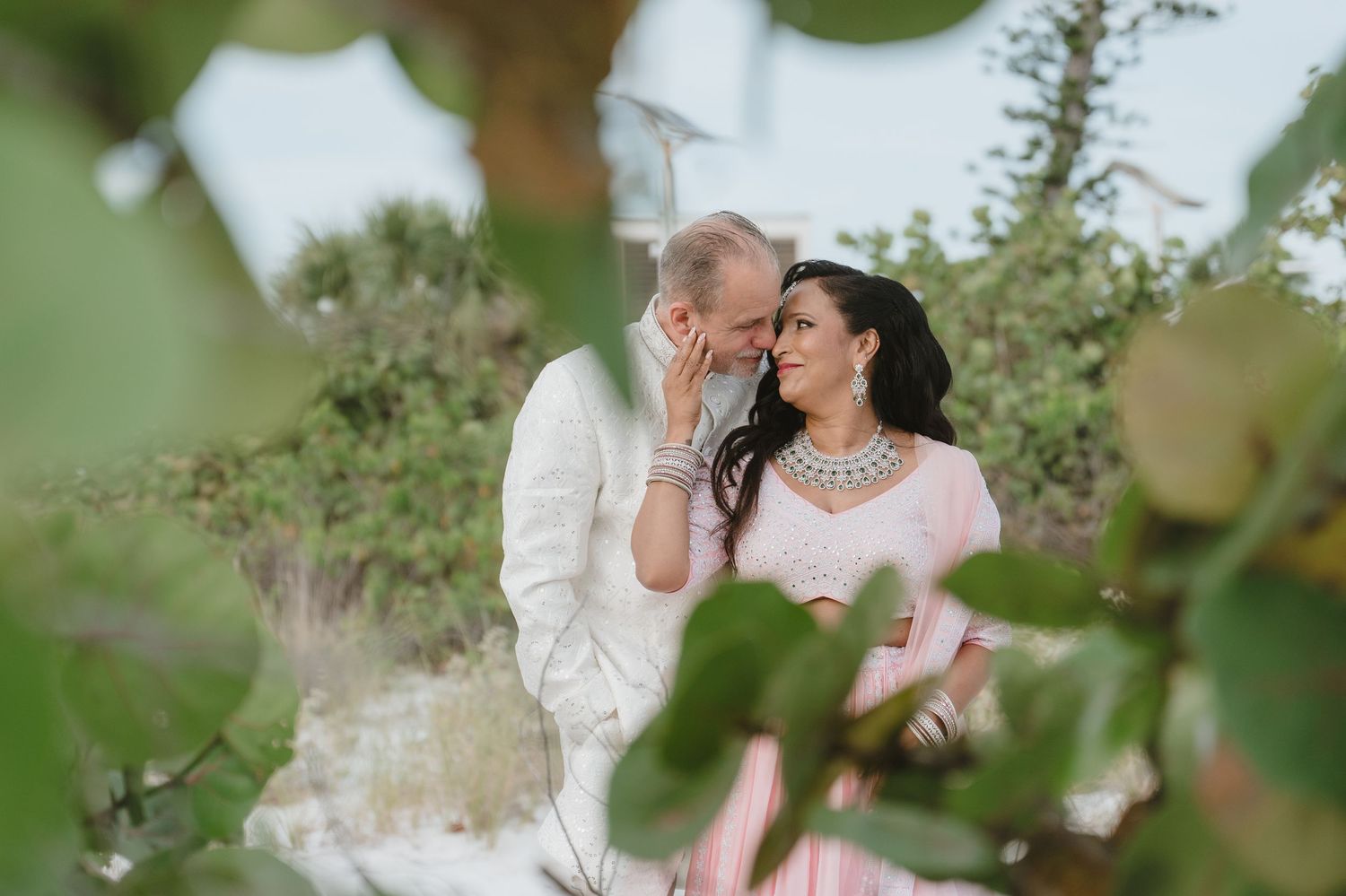 Couple in elegant attire sharing a romantic moment outdoors, framed by lush green foliage in a serene natural setting.