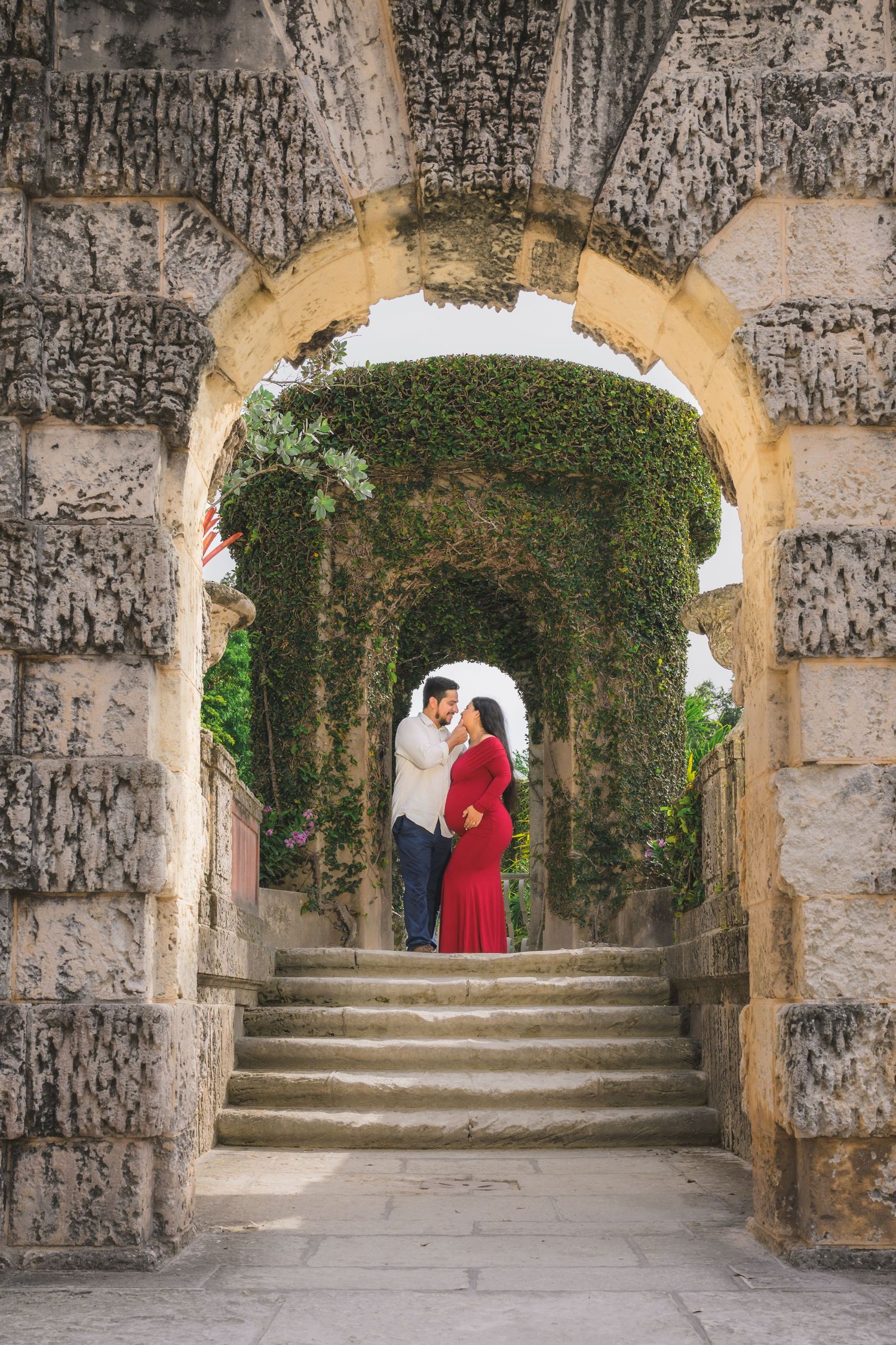 Couple embracing under stone arches covered in ivy, standing on steps in a romantic garden setting.