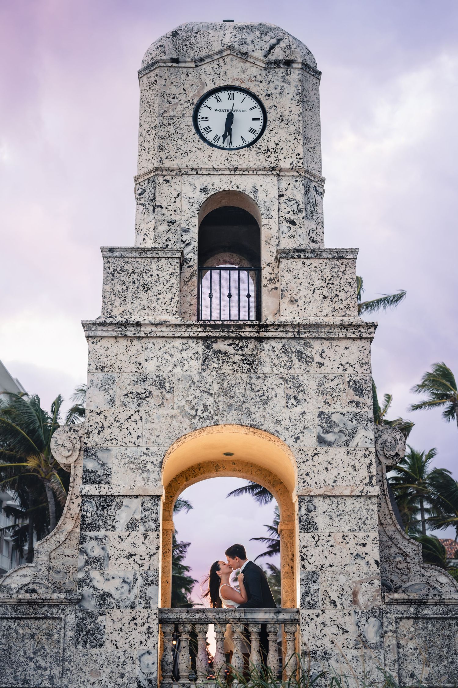 Couple sharing a romantic kiss under a historic clock tower arch at dusk.