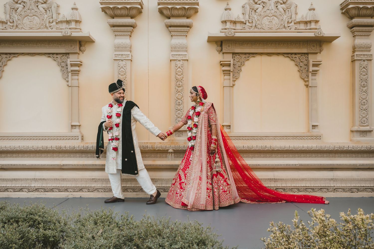 Indian bride and groom in traditional red and black attire hold hands at ornate venue.