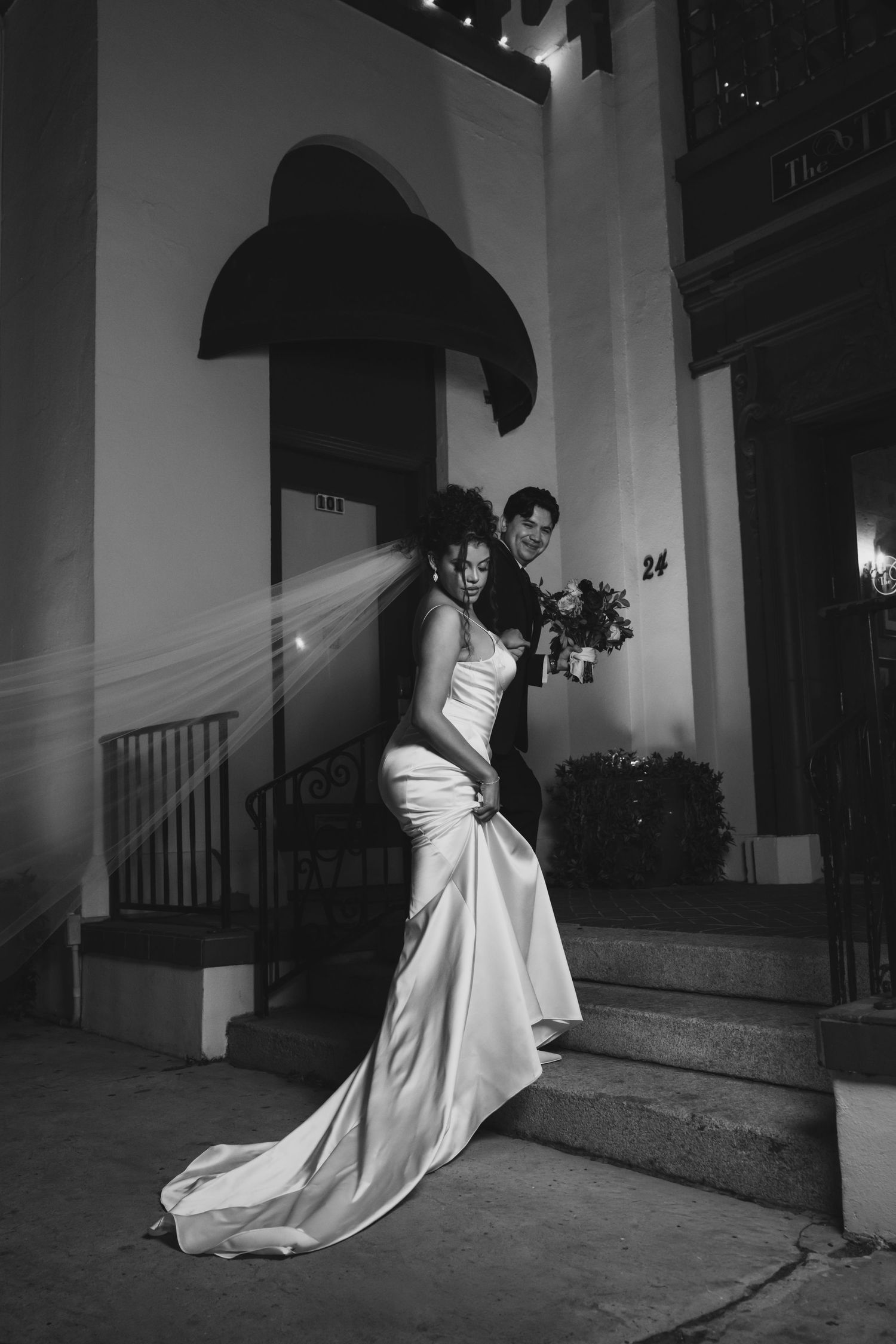 Black and white wedding photo of bride in elegant gown on steps with groom holding bouquet at night.