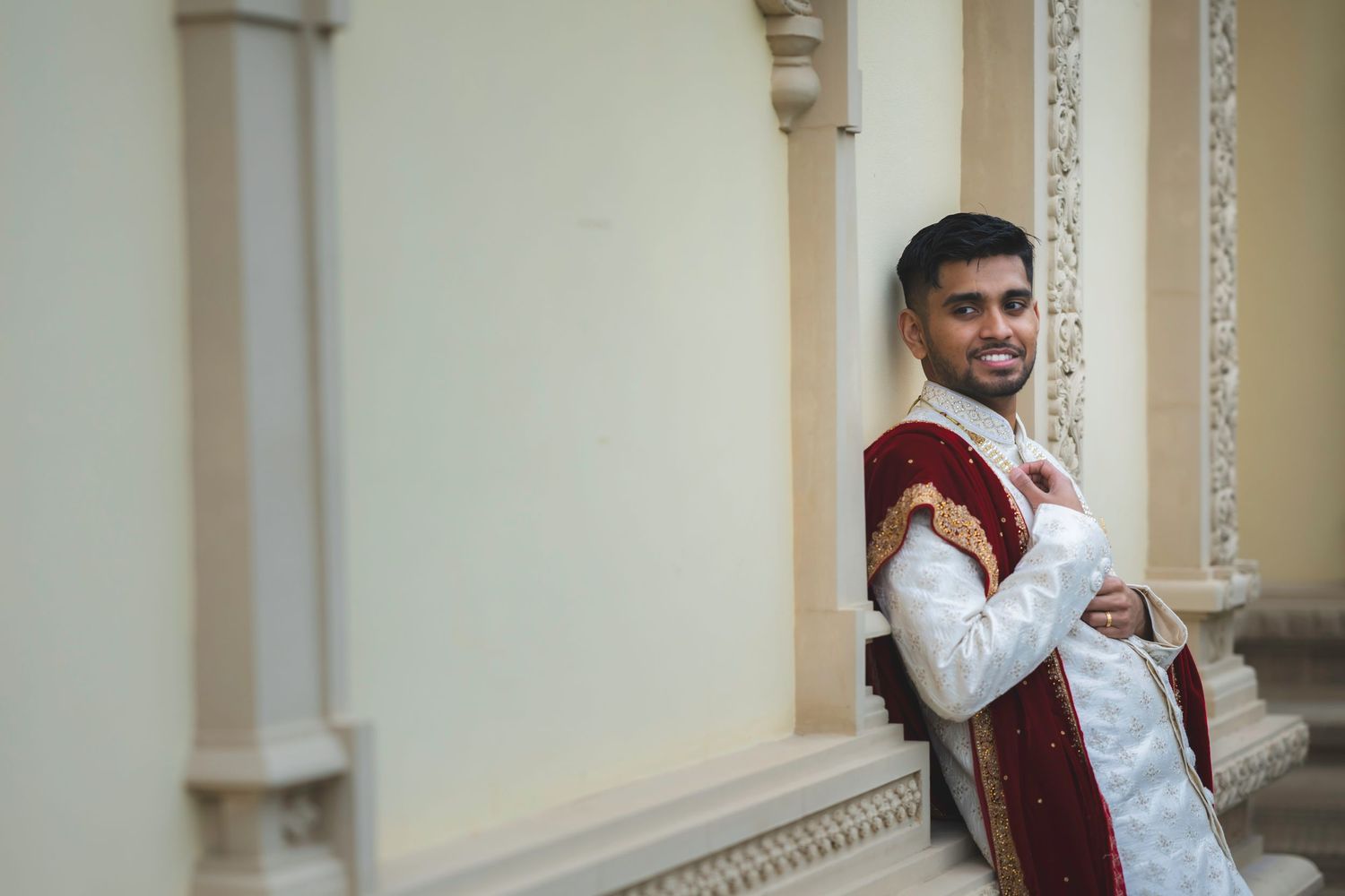 Indian groom in white sherwani with red dupatta smiles while leaning against decorative white columns.