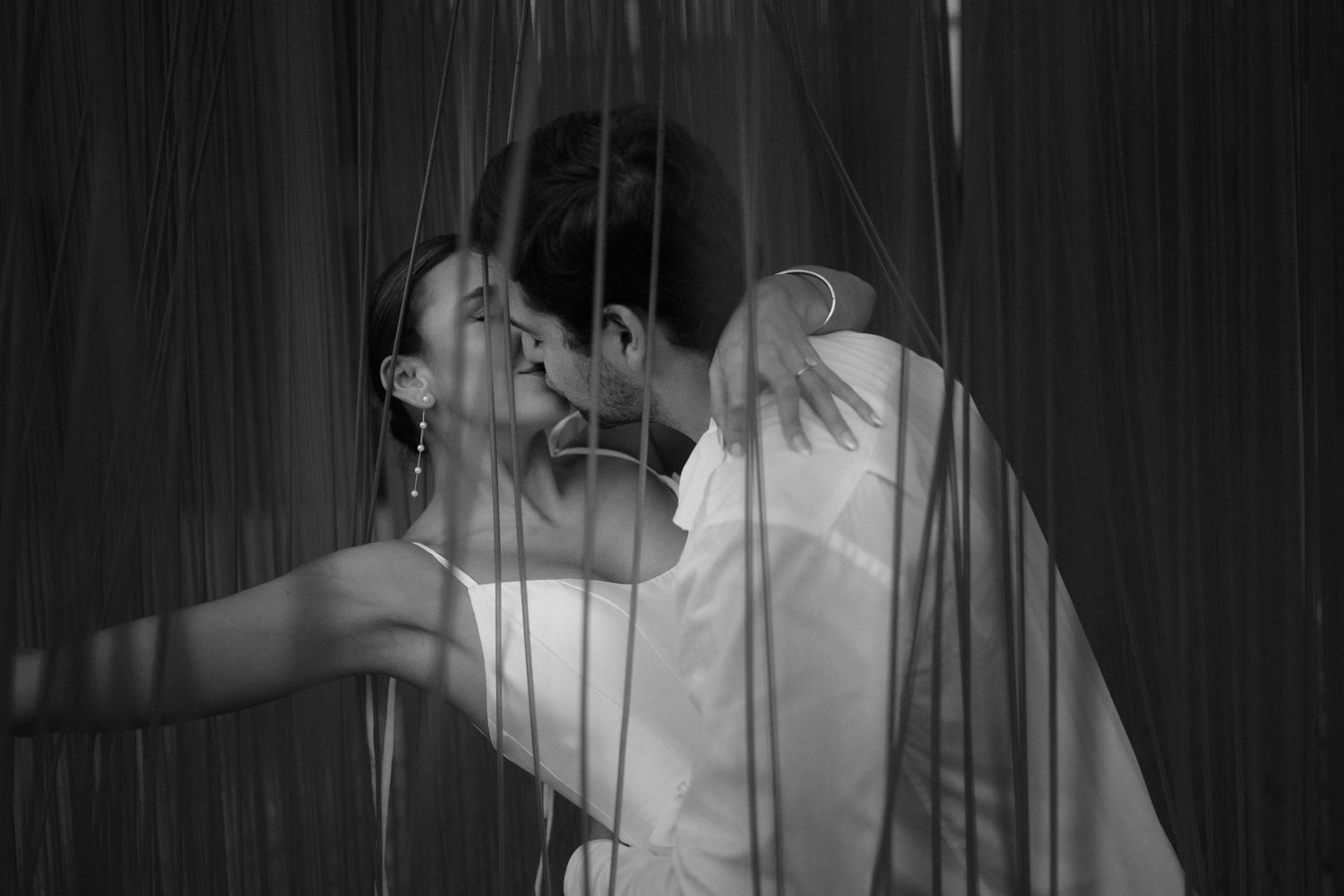 Romantic black and white photo of a couple sharing a kiss through hanging metal string curtains.