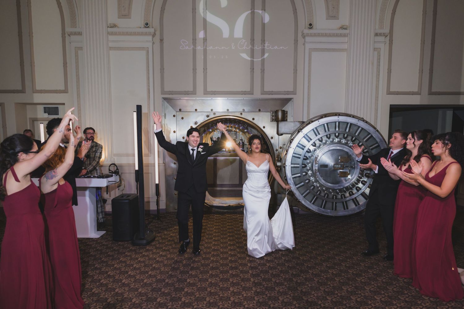 Bride and groom make grand entrance at wedding reception with bridesmaids in burgundy dresses cheering beside a large vault door.