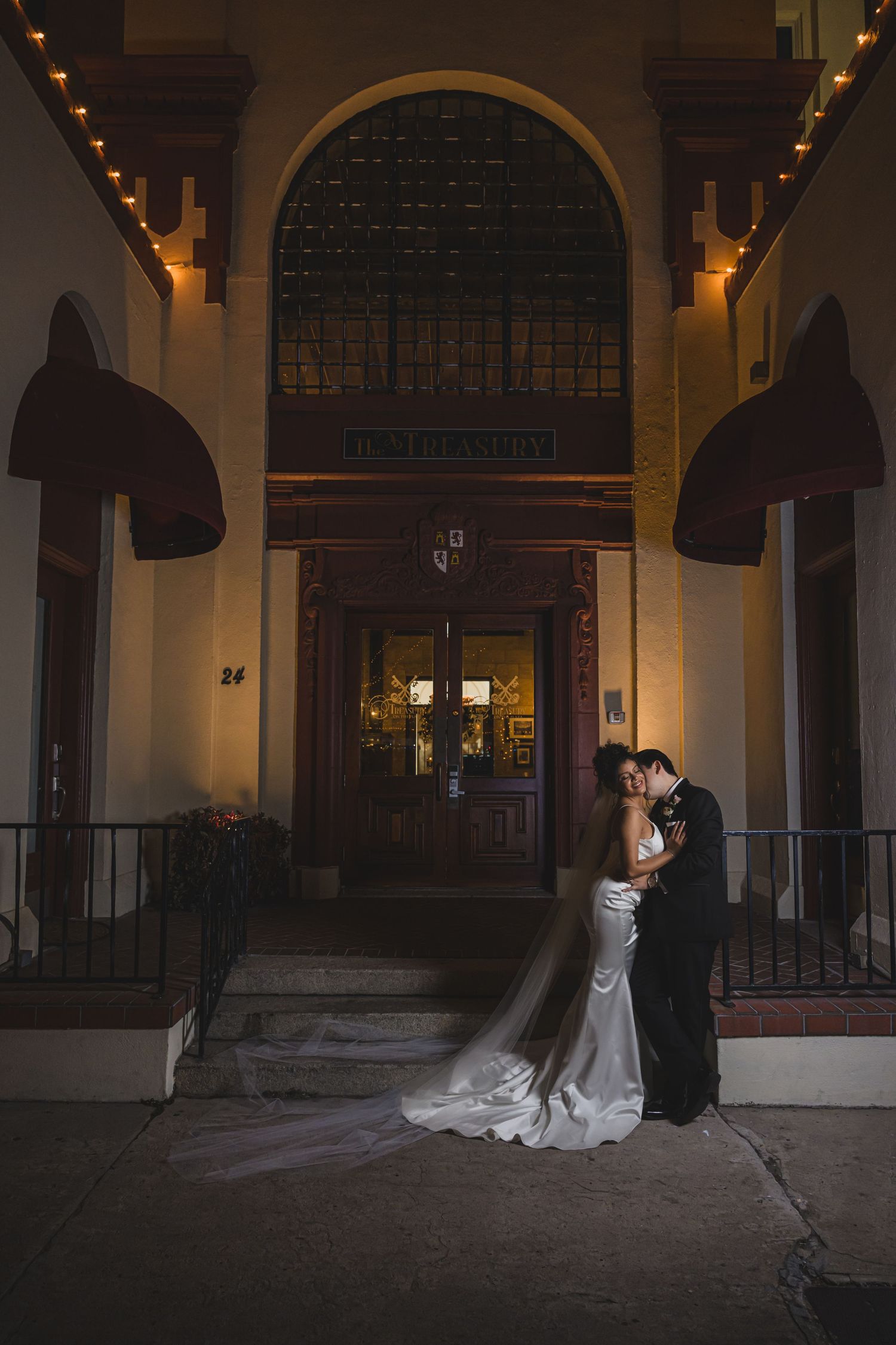 Bride and groom share a romantic kiss at night outside an elegant venue entrance with warm string lights.