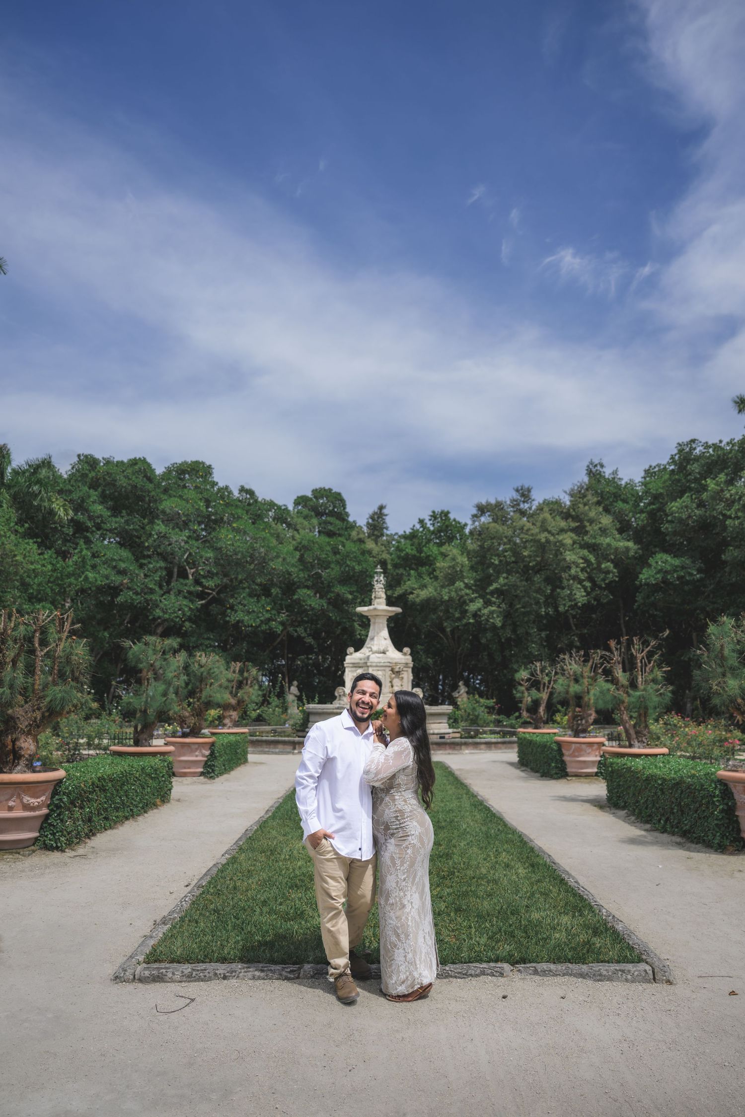 Couple embracing in a formal garden with fountain, lush trees, and blue sky backdrop.