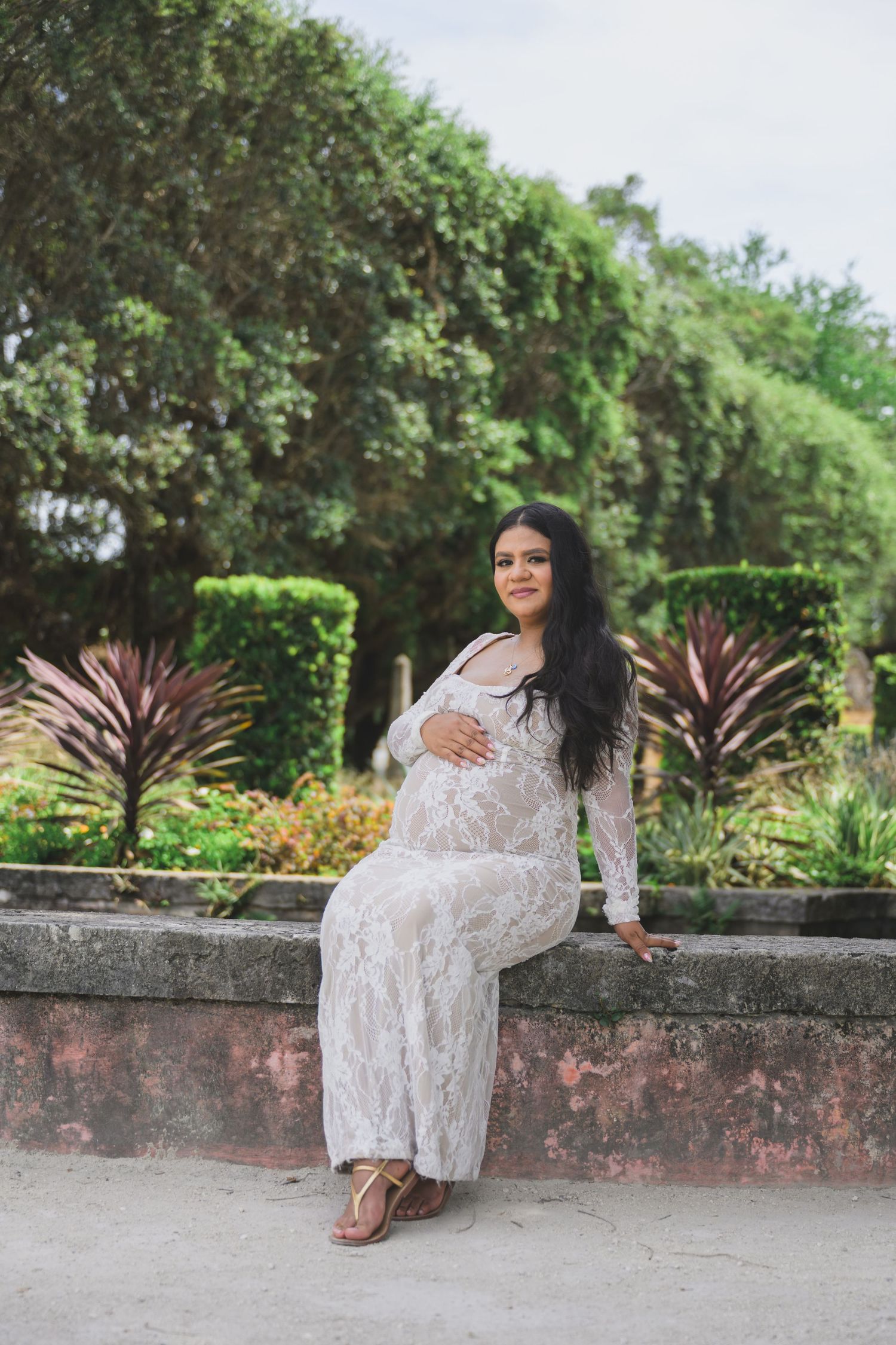 Pregnant woman in white lace dress sitting on stone wall in lush green garden, cradling baby bump.