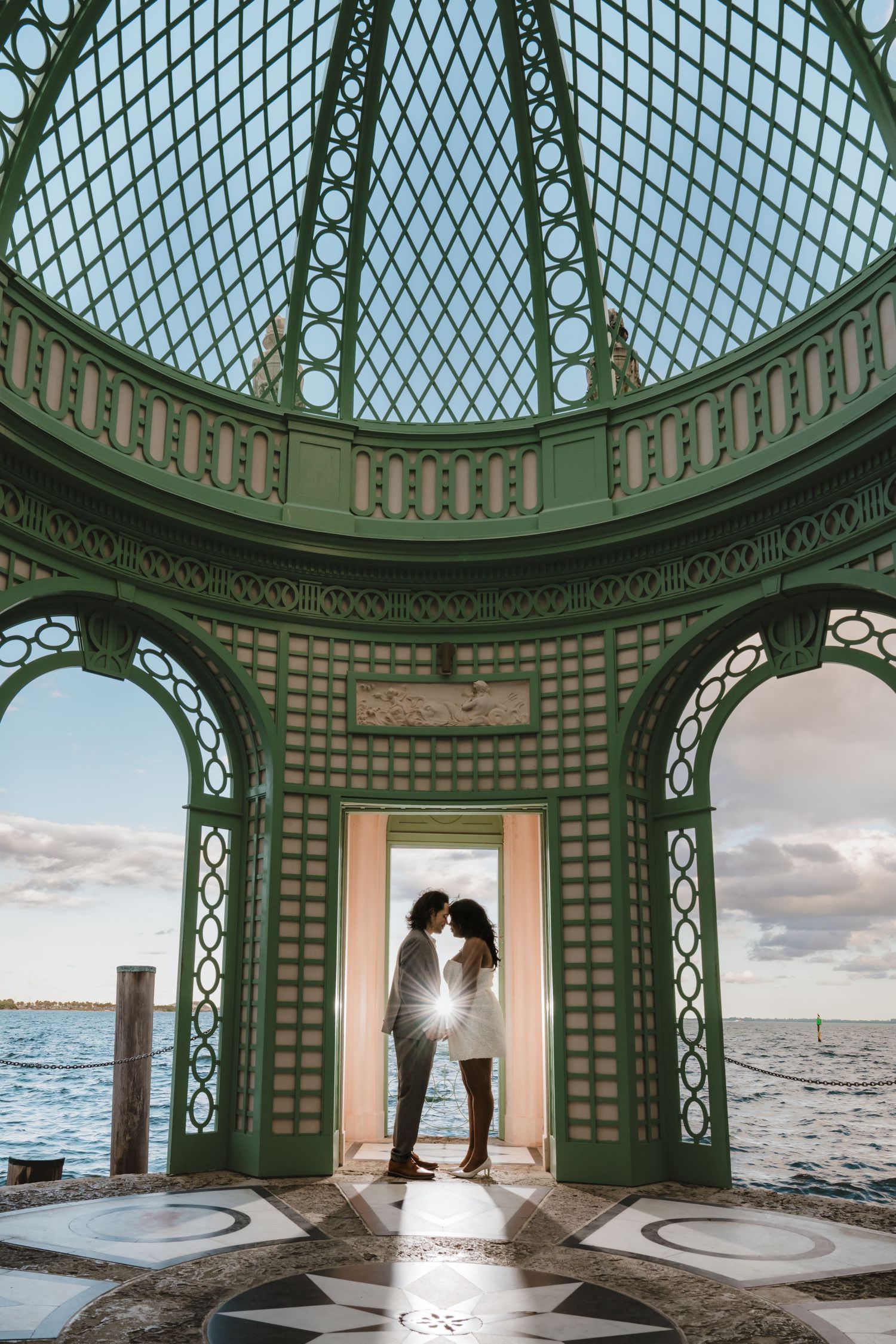 Couple sharing a romantic moment in an ornate green gazebo overlooking the ocean at sunset.