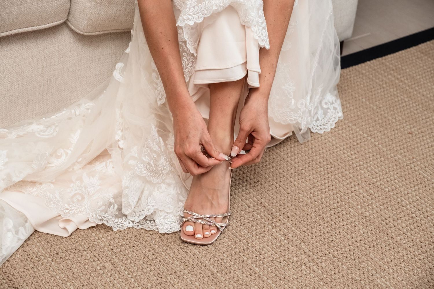 Bride in lace wedding gown fastening strappy heeled sandal while seated on beige sofa.