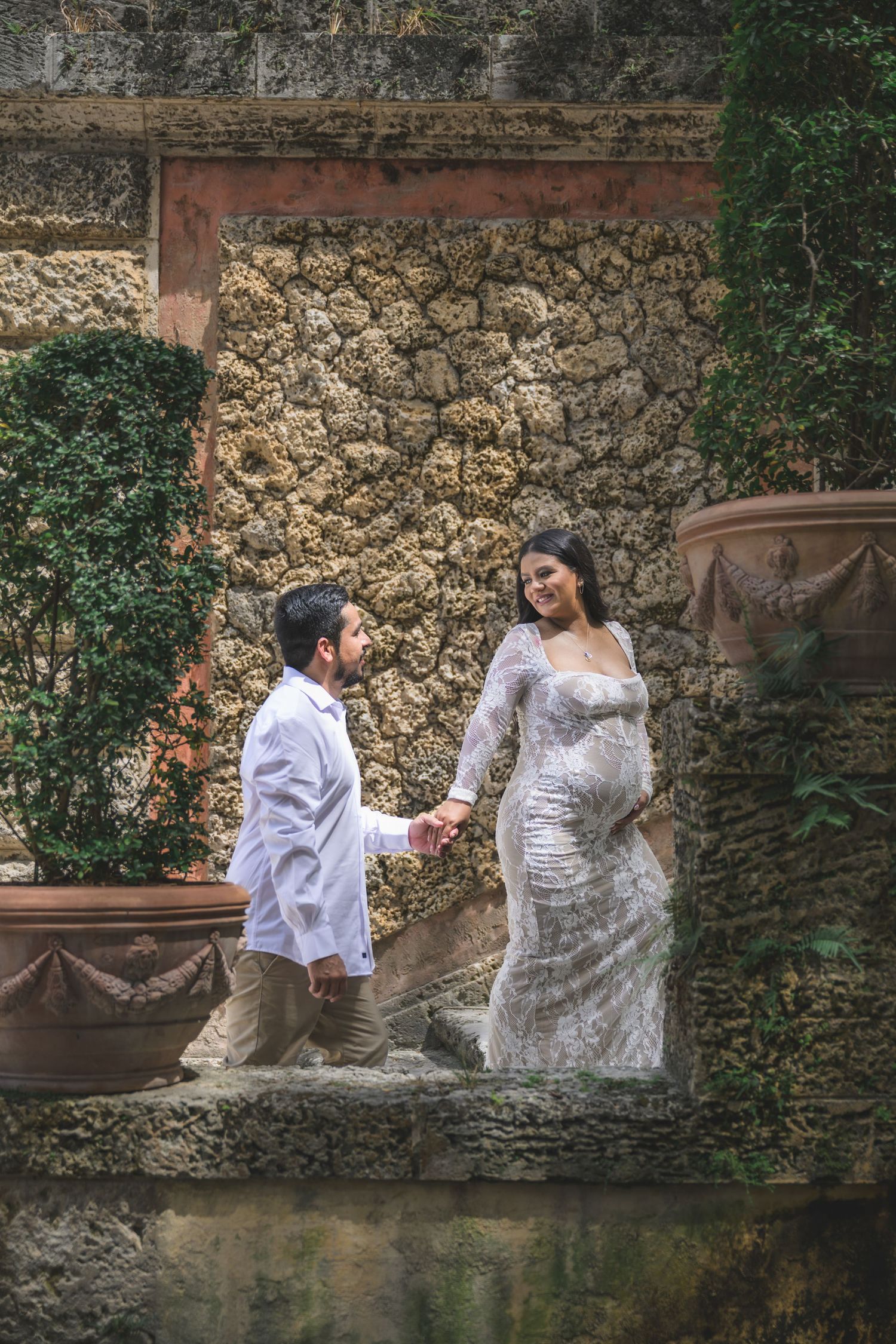 Pregnant woman in white lace gown holds hands with man in garden with stone wall and lush greenery.