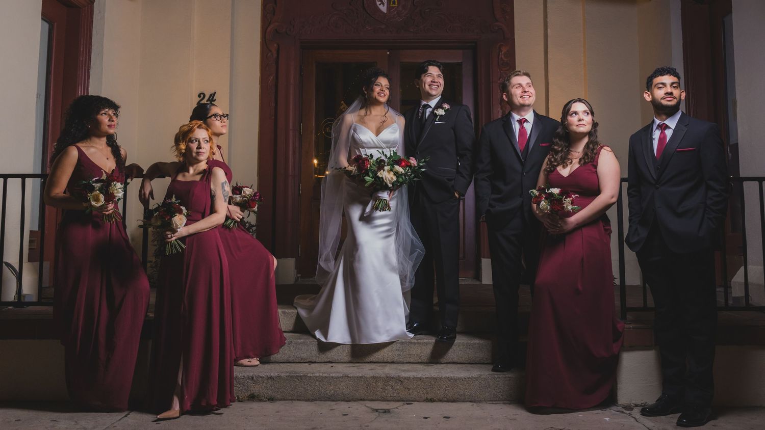 Wedding party poses on steps, bride in white gown, bridesmaids in burgundy dresses, groomsmen in dark suits.
