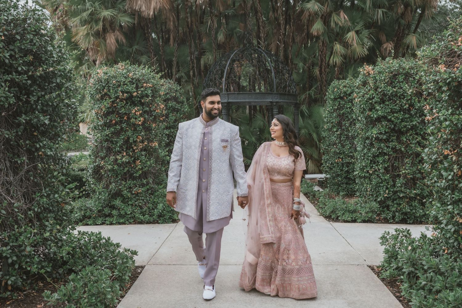 Indian bride and groom in traditional attire walking hand in hand along a garden pathway lined with hedges.