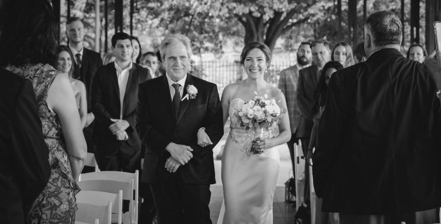Bride walking down the aisle with her father in a black and white outdoor wedding ceremony photo.
