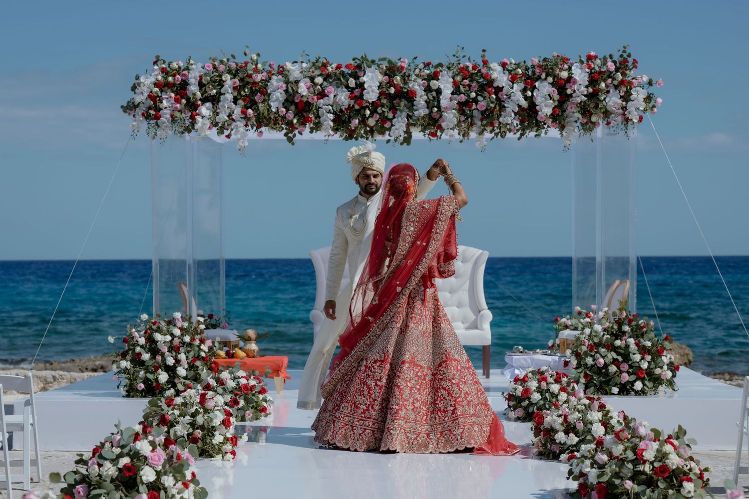 Indian bride and groom stand under floral arch at beachside wedding ceremony with ocean backdrop.