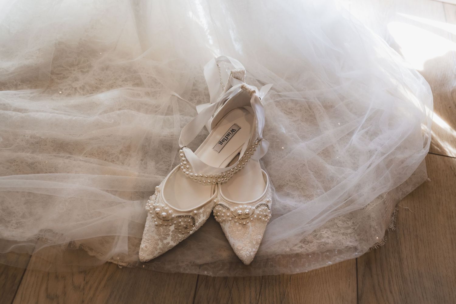 Pearl-embellished white bridal flats resting on flowing tulle wedding dress on wooden floor.