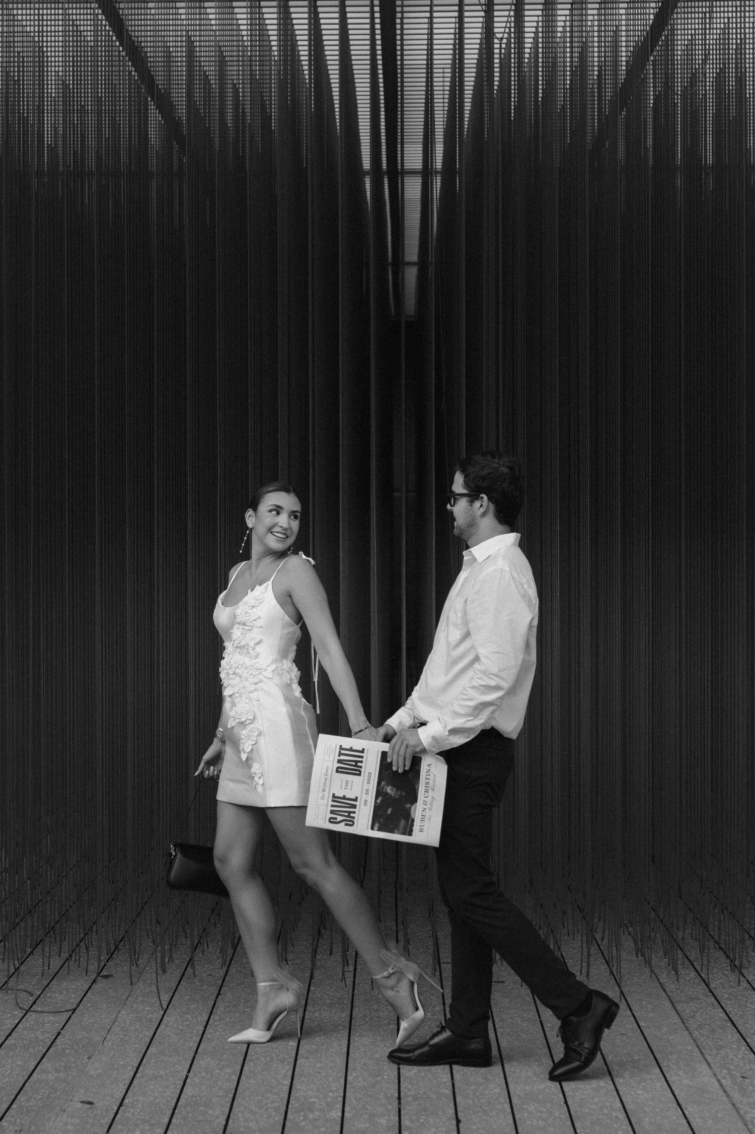 Black and white photo of a stylish couple holding hands and walking past a striped metal wall.
