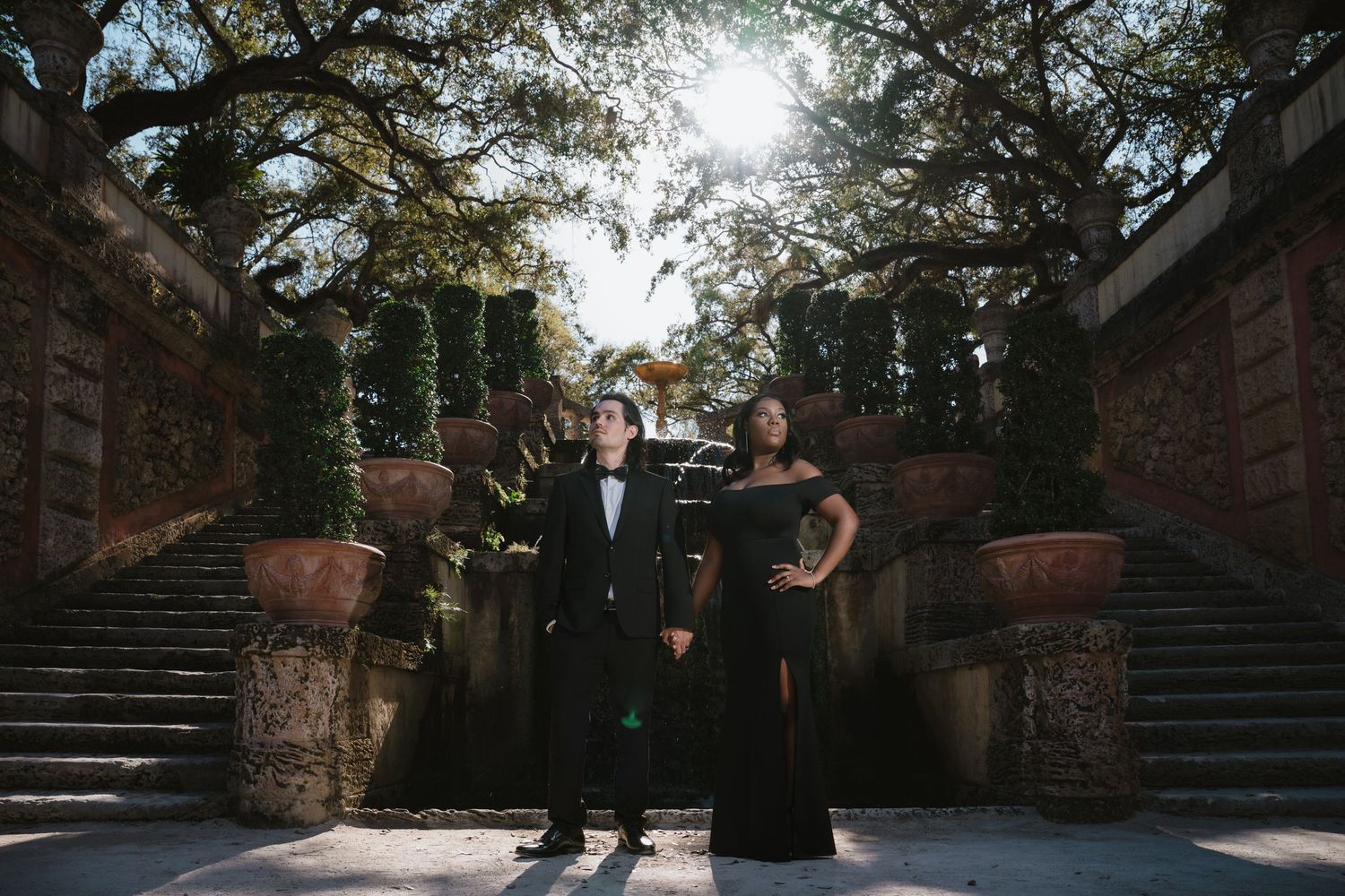 Couple in black formal wear standing together in sunlit garden with grand stone staircase.