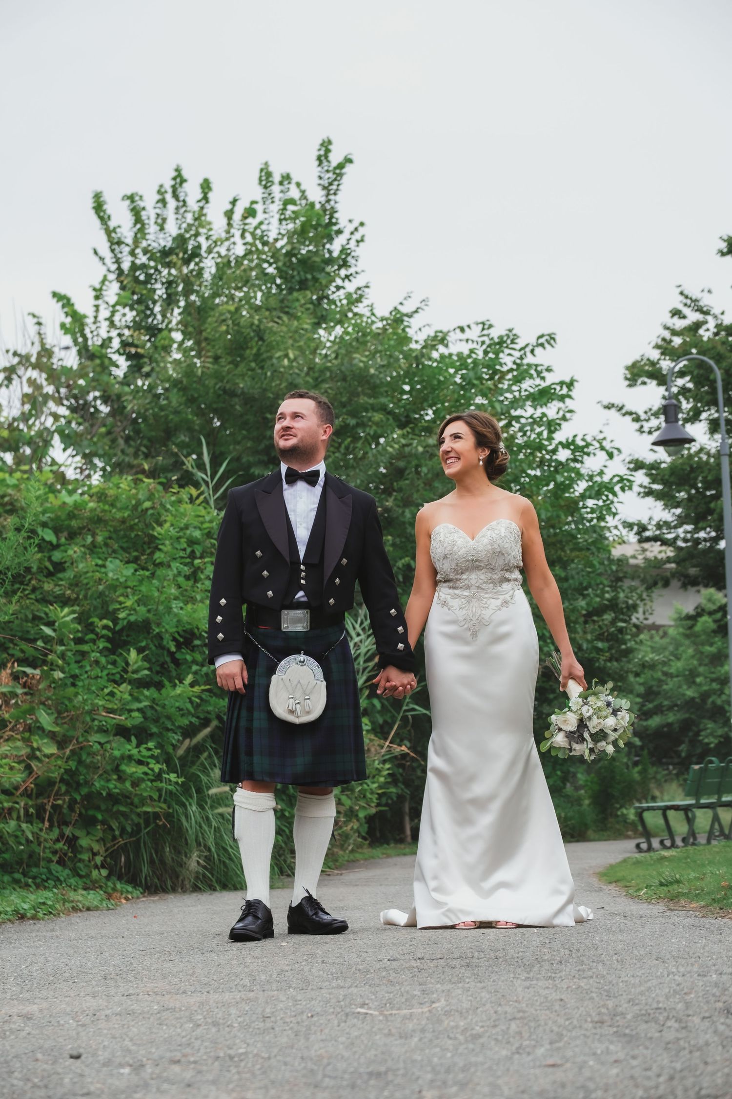 Groom in Scottish kilt and bride in white gown walking hand in hand through lush garden path.