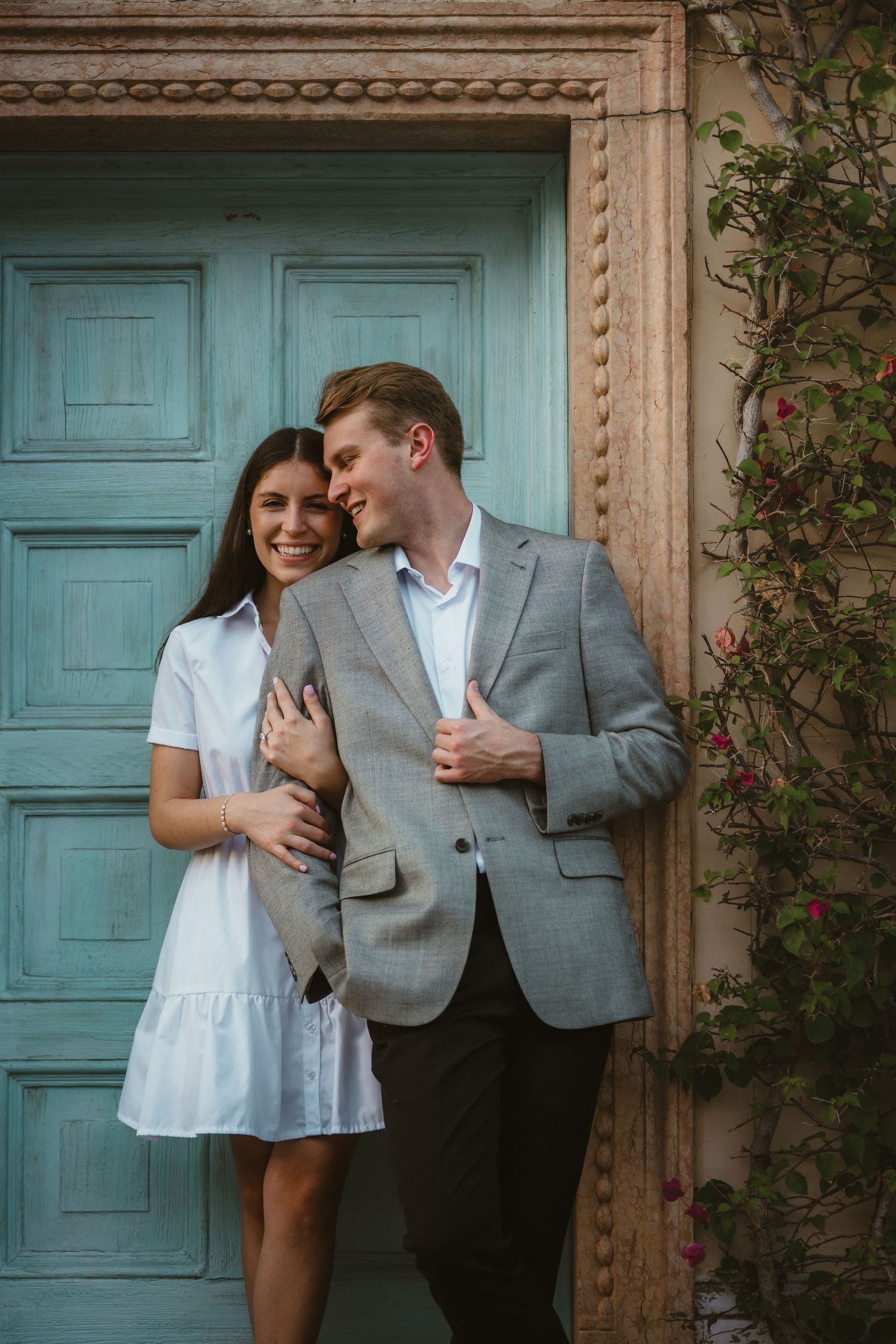 Couple embracing and smiling in front of a turquoise door with climbing flowers.