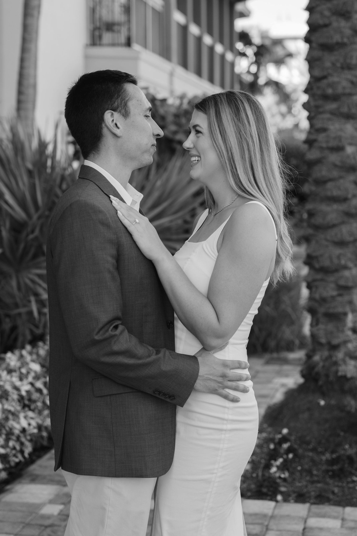 Black and white photo of a couple embracing and smiling at each other outdoors near tropical plants.