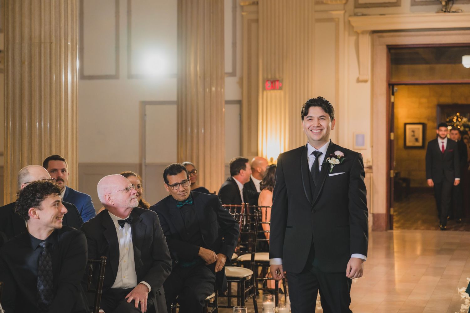 Groom in black tuxedo smiling at wedding ceremony aisle with seated guests in elegant venue.