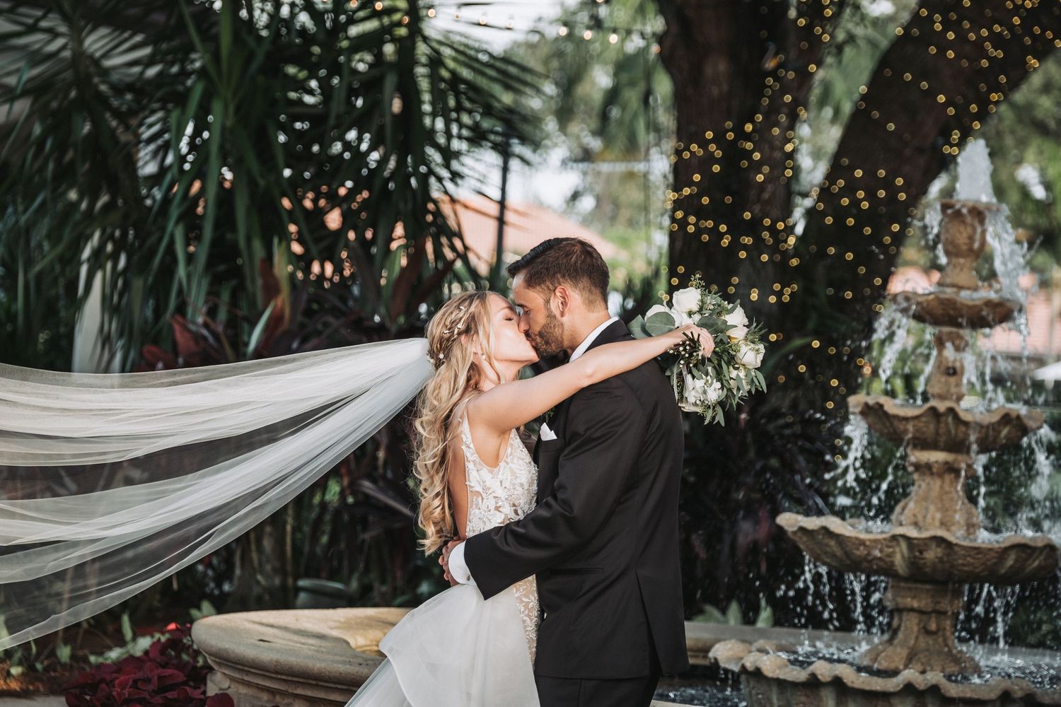 Bride and groom share a romantic kiss near a stone fountain, surrounded by lush greenery and twinkling fairy lights.