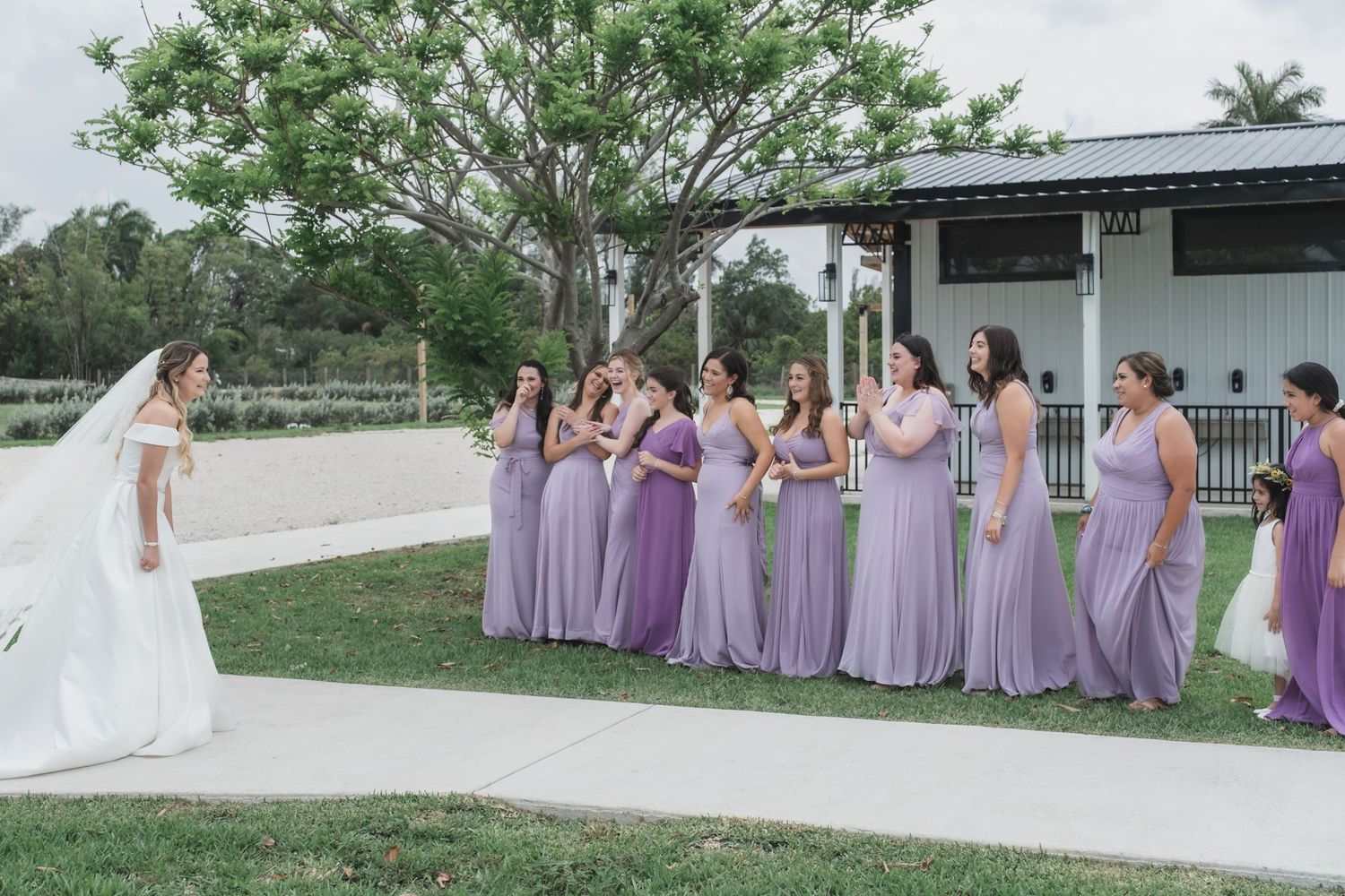 Bride in white gown reveals herself to bridesmaids in matching lavender dresses outdoors.