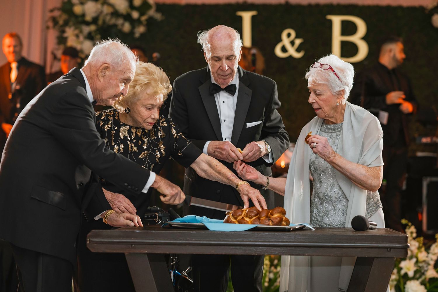 Four elderly guests cutting challah bread at a Jewish wedding reception under monogram letters J and B.