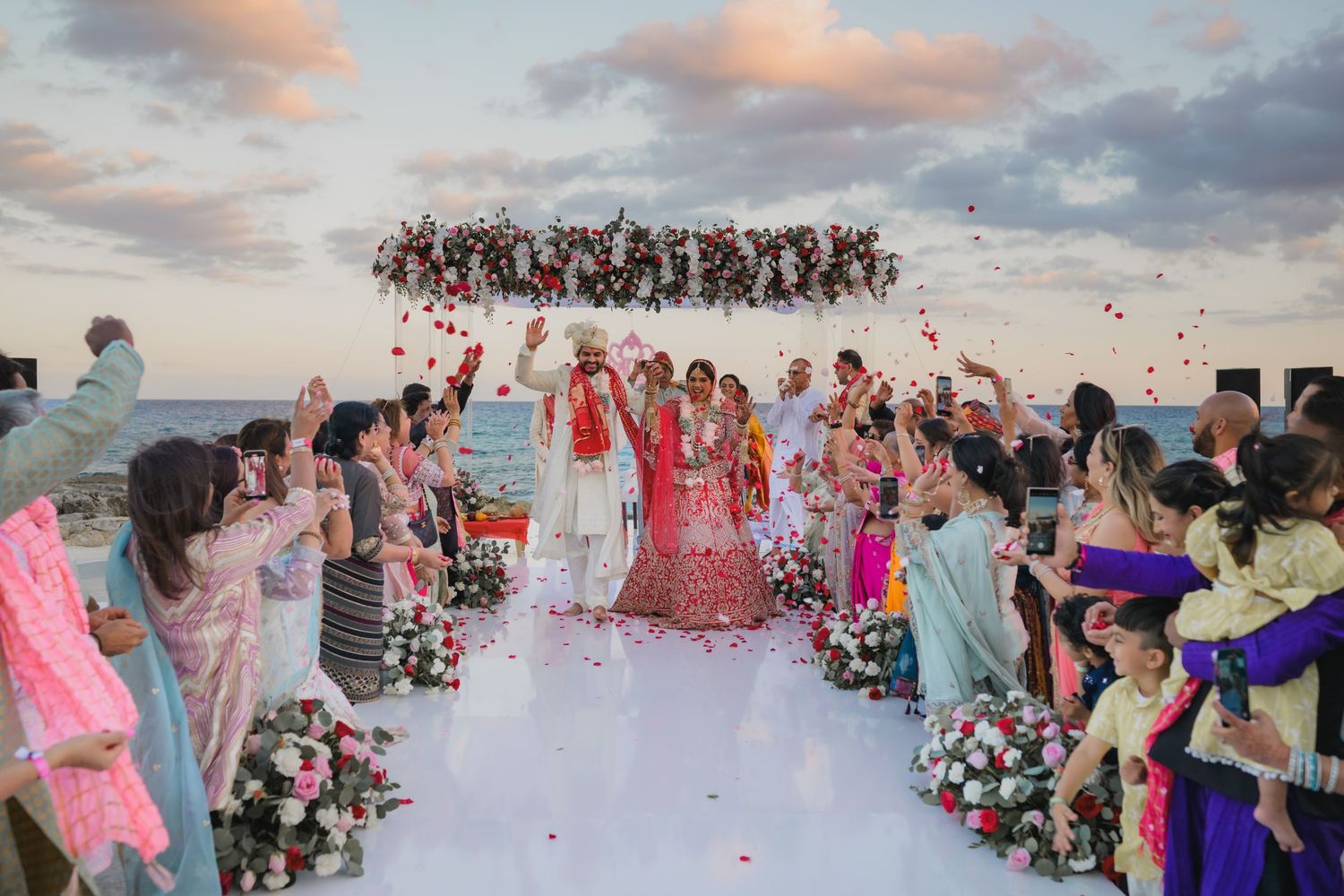 Indian wedding couple celebrate under floral mandap as guests toss rose petals at sunset beach.